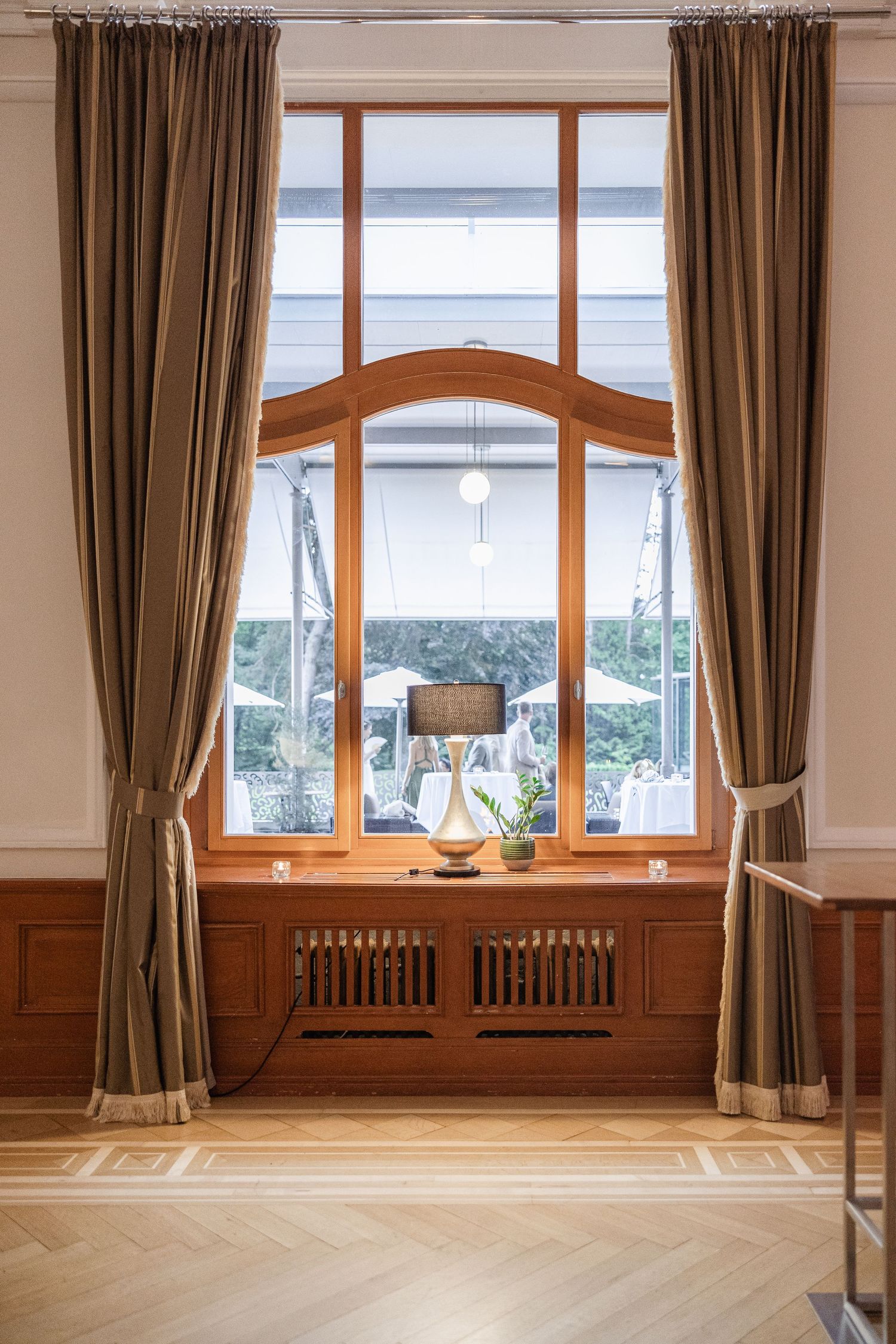 Large empty room with hardwood floors, tall windows with brown curtains, and wooden wainscoting along the walls.