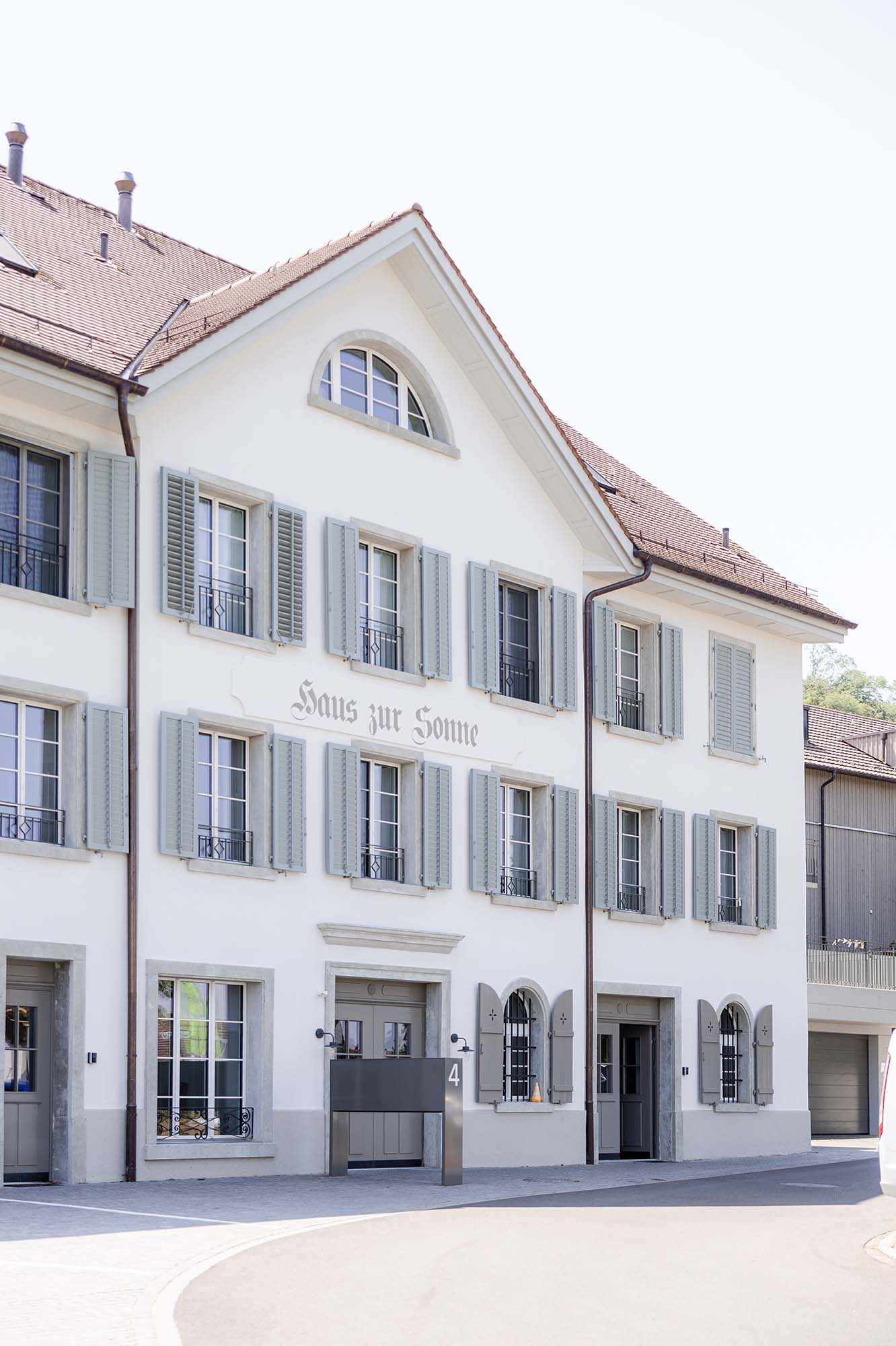 White Swiss building with green shutters and red roof, featuring the inscription Haus zur Sonne on its facade.