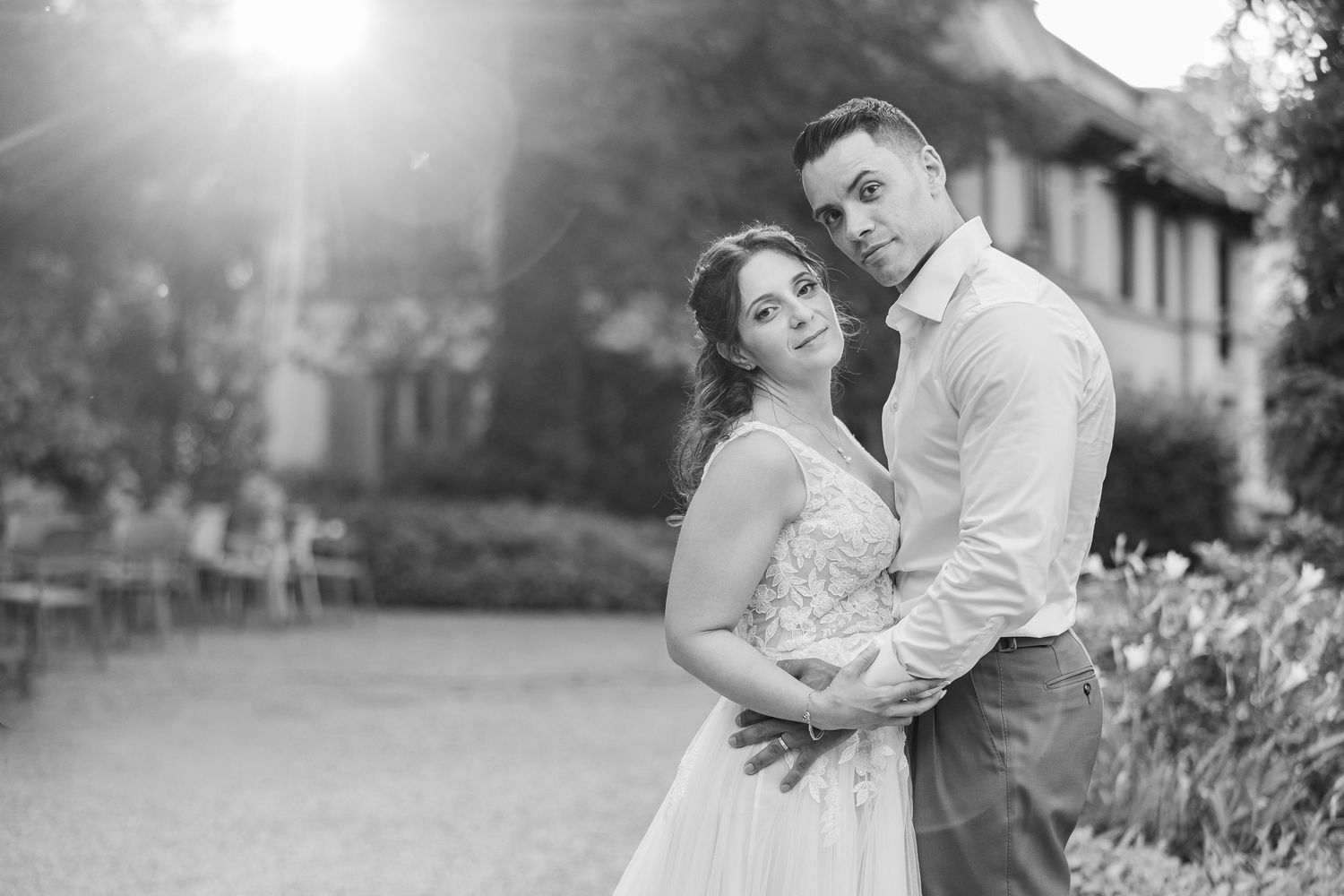 Black and white photo of a couple embracing outdoors at a wedding venue with soft sunlight in the background.