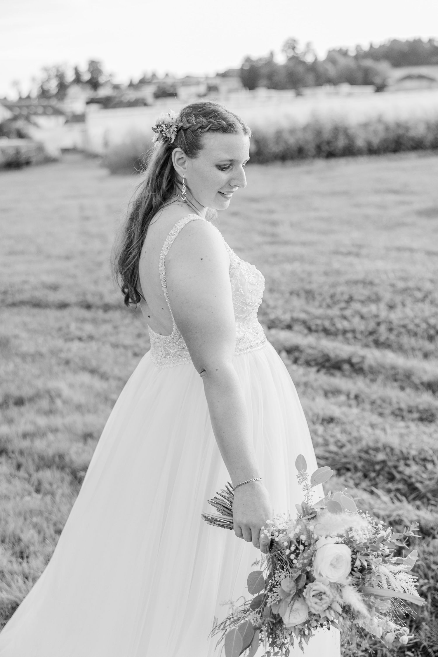 A black and white side profile of a bride in a flowing lace wedding gown holding a floral bouquet in a field.