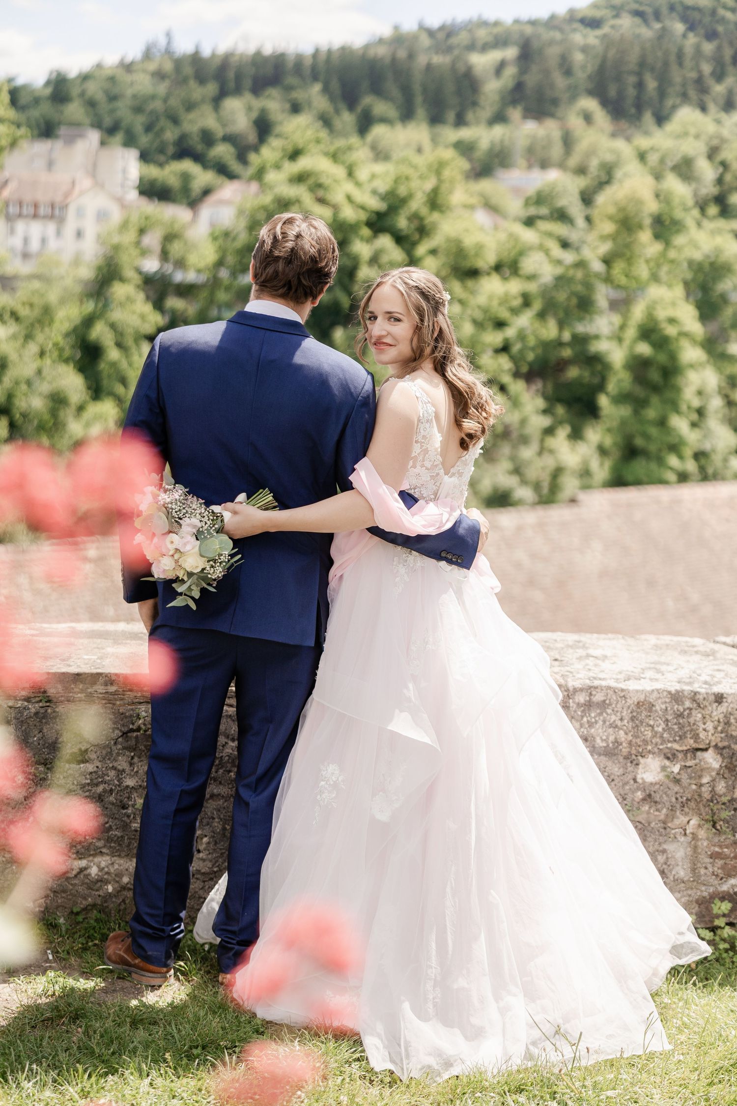 A romantic wedding couple embraces while standing on a stone wall overlooking a scenic green countryside landscape.