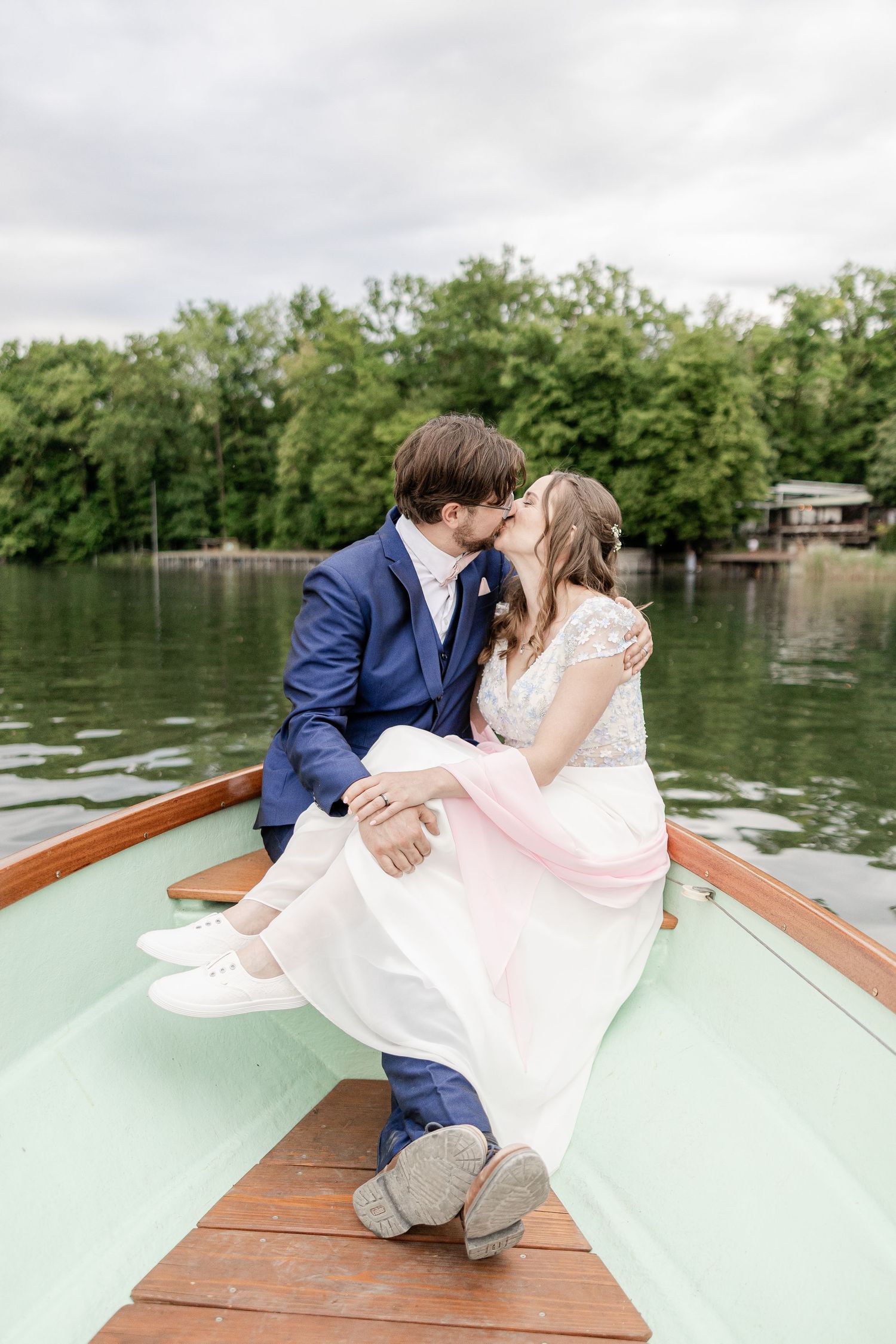 A romantic couple shares a tender moment while sitting in a wooden boat on a serene lake surrounded by trees.