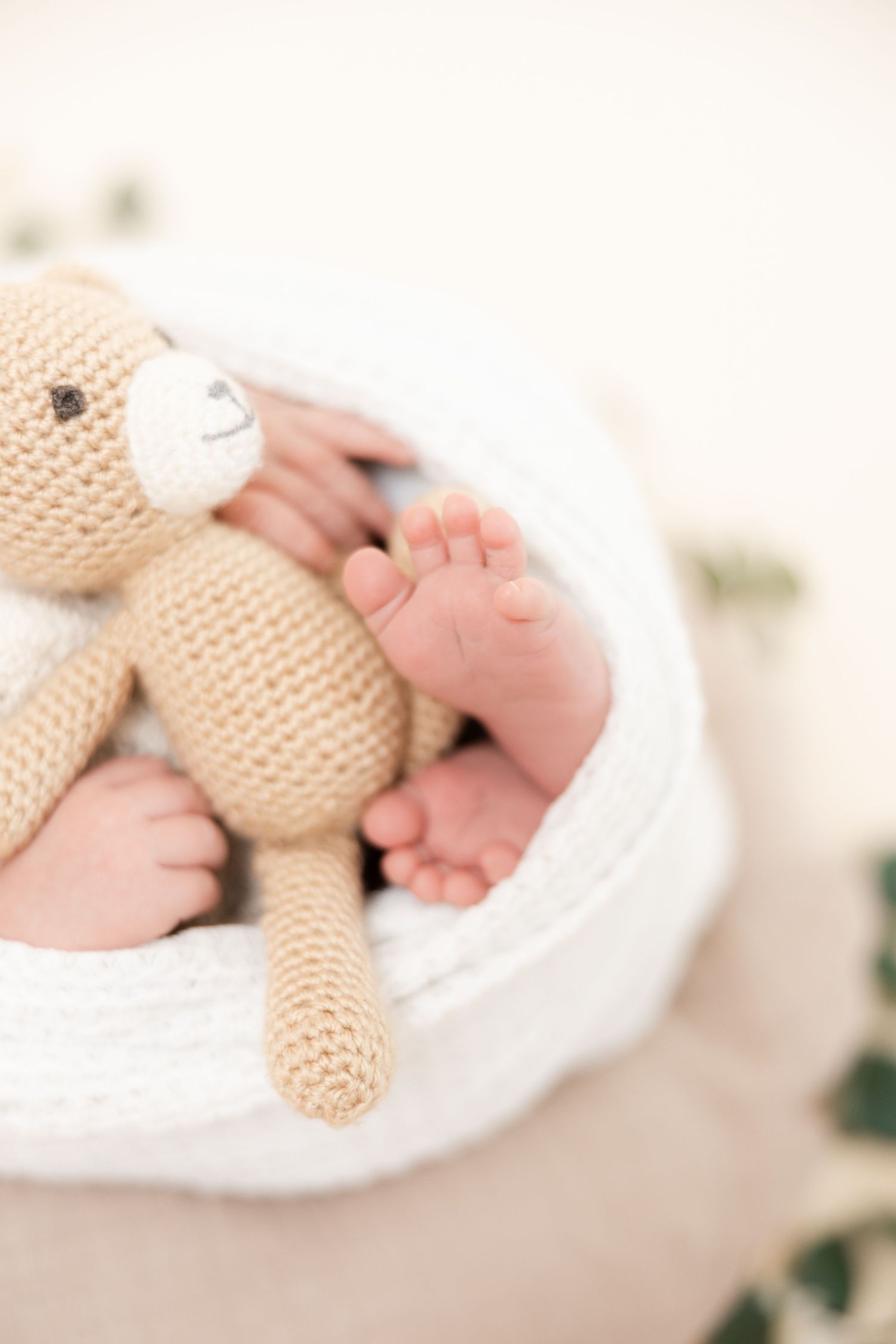 Baby's tiny feet rest on white fabric next to a small knitted teddy bear in soft neutral tones.