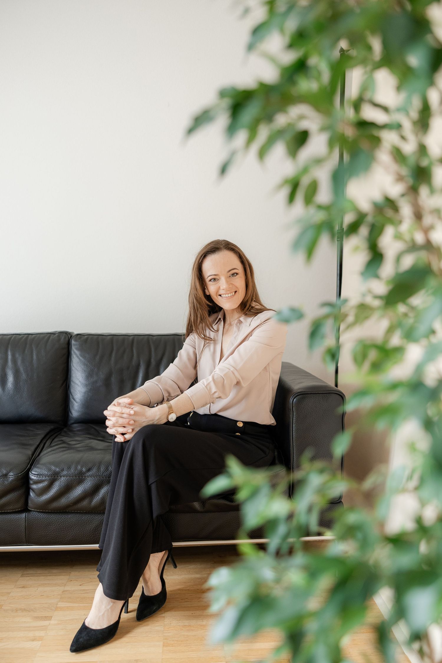 Someone relaxes on a black leather couch wearing a pink blouse and black pants, with green plants in the foreground.