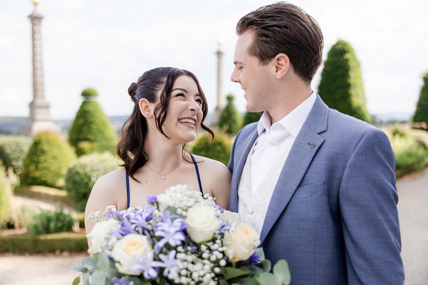 Smiling couple in formal attire holding white and purple bouquet in a manicured garden with topiaries.