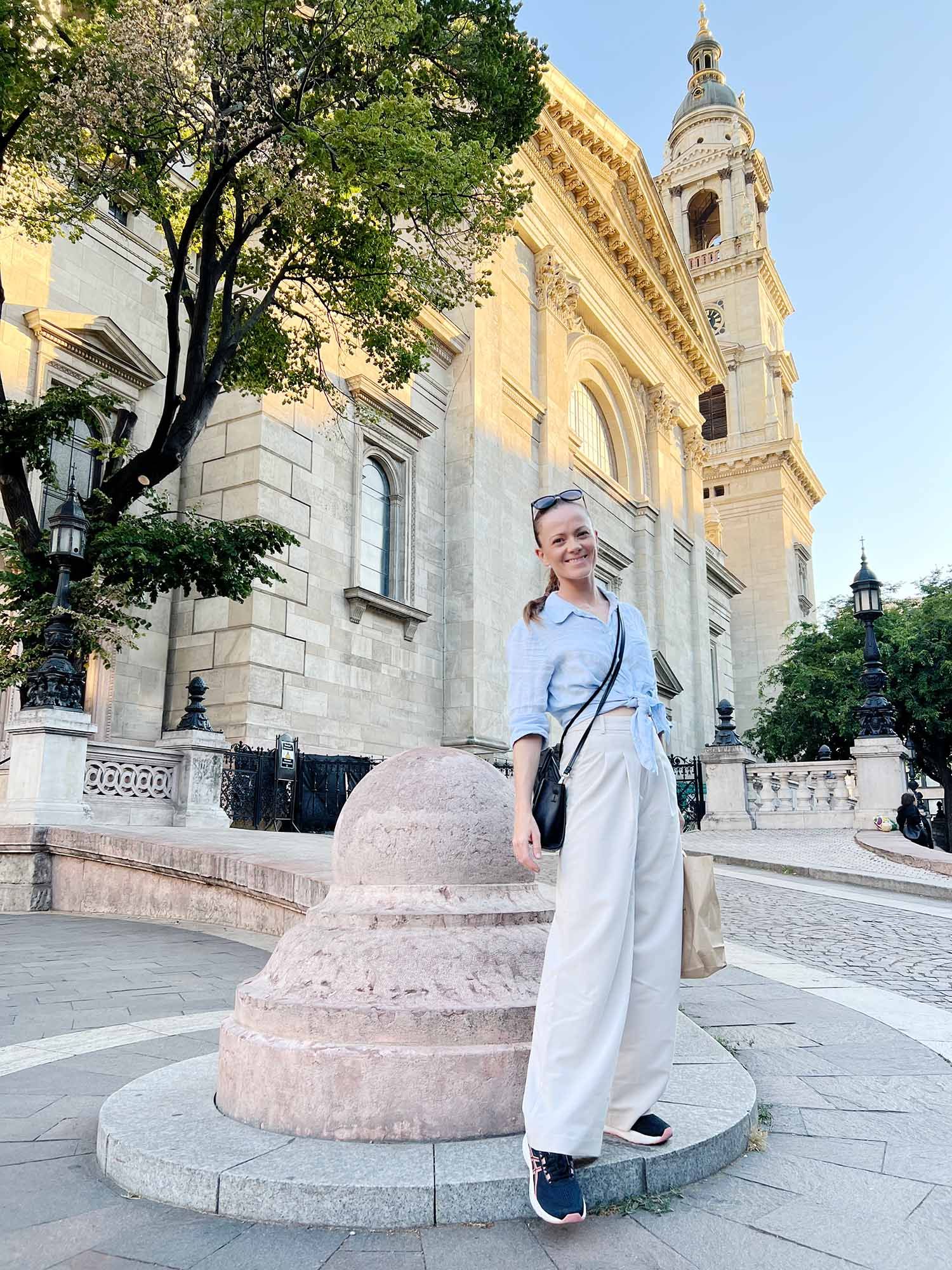 A person stands next to a stone bollard outside the ornate St. Stephen's Basilica in Budapest on a sunny day.