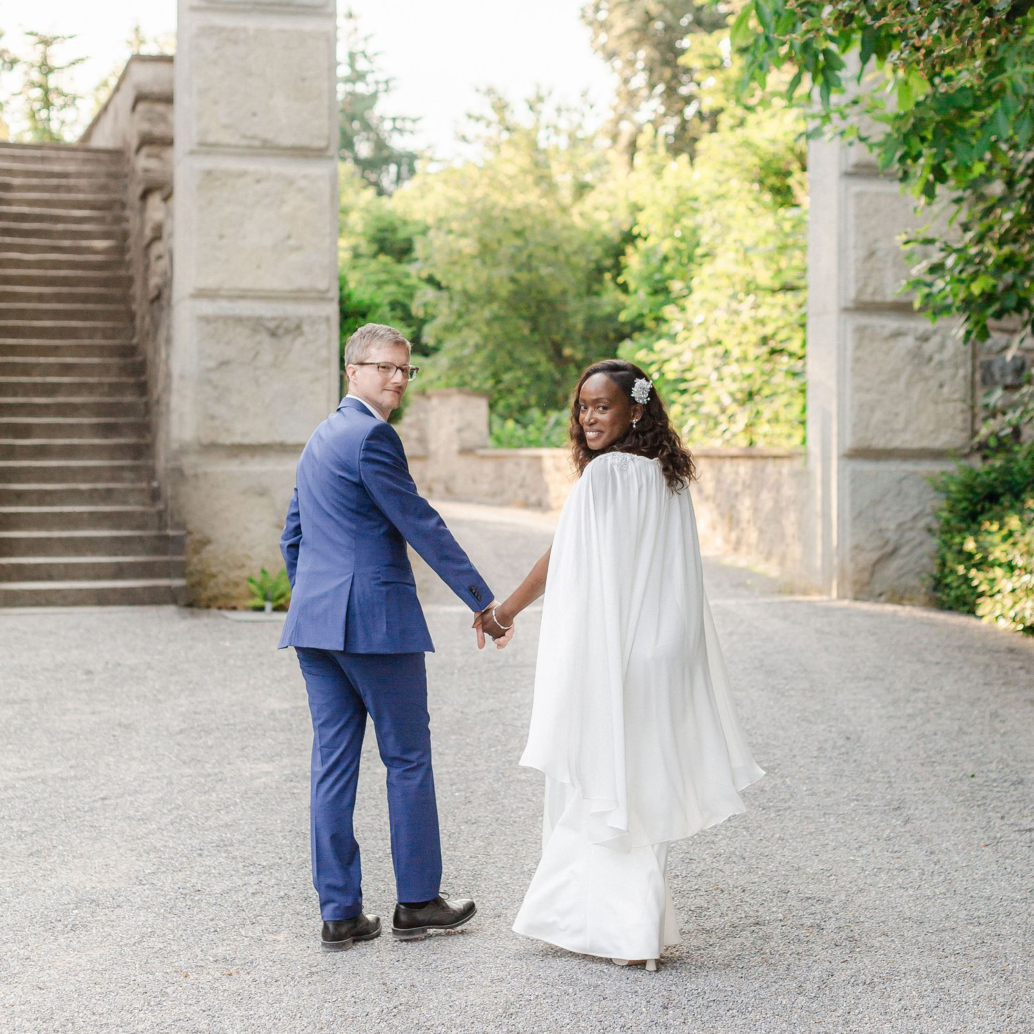 A couple in formal attire walks together on a stone path surrounded by greenery and architectural elements.