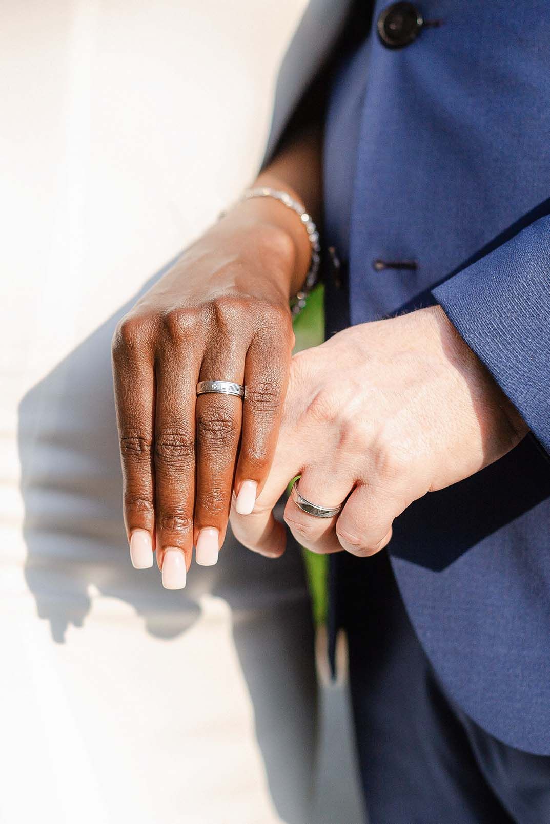 Close-up of two hands wearing wedding bands and a navy blue suit jacket showing an intimate wedding moment.