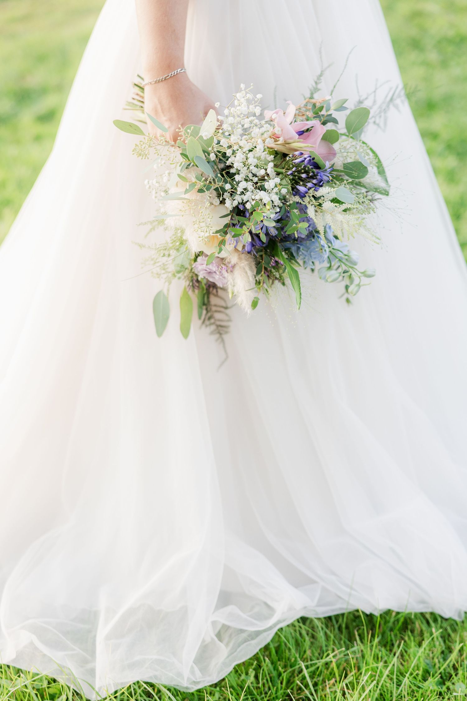 A cascading white wedding dress hem with delicate floral bouquets featuring purple and white blooms against lush grass.