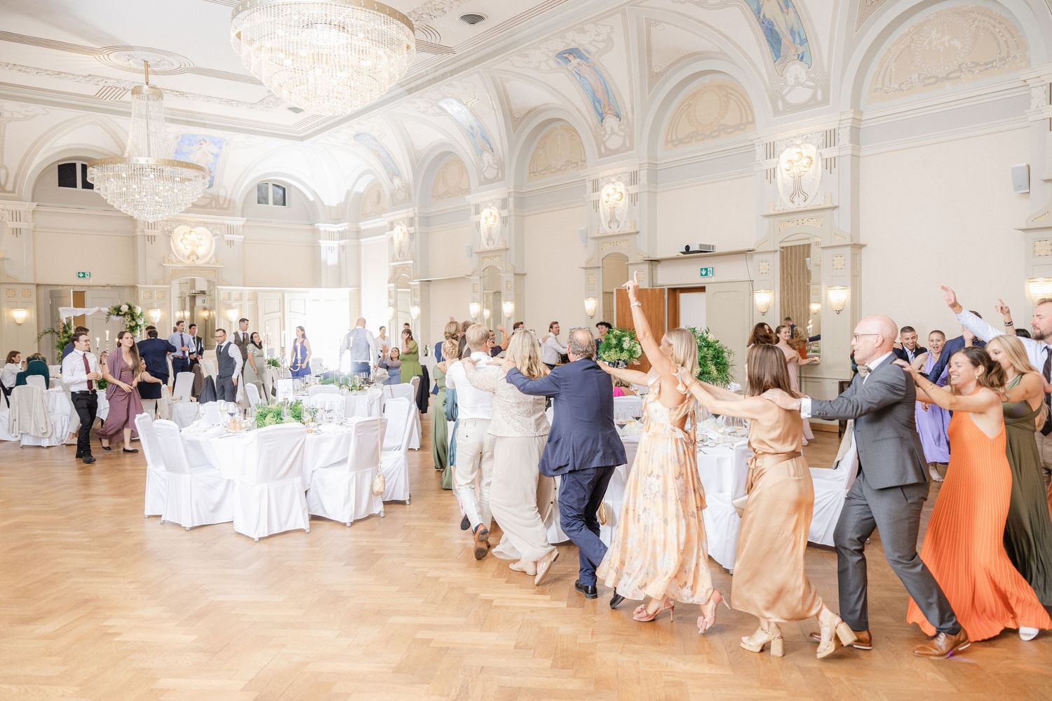 Wedding party dances in a line through an elegant white ballroom with ornate architectural details and chandeliers.