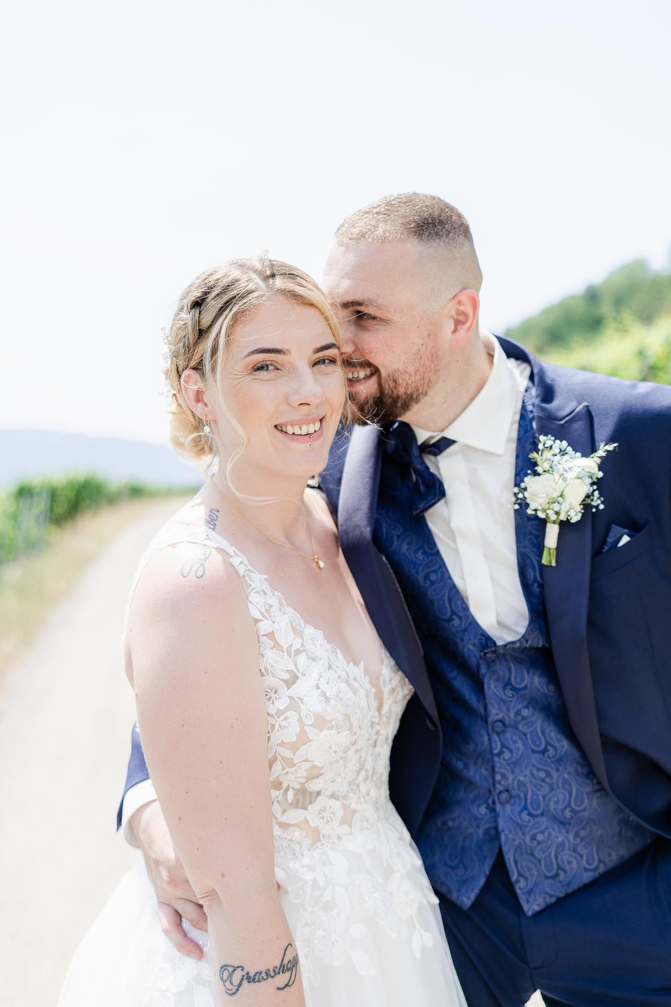 A smiling bride in a lace gown poses with a groom in a navy blue suit on a sunny outdoor path.