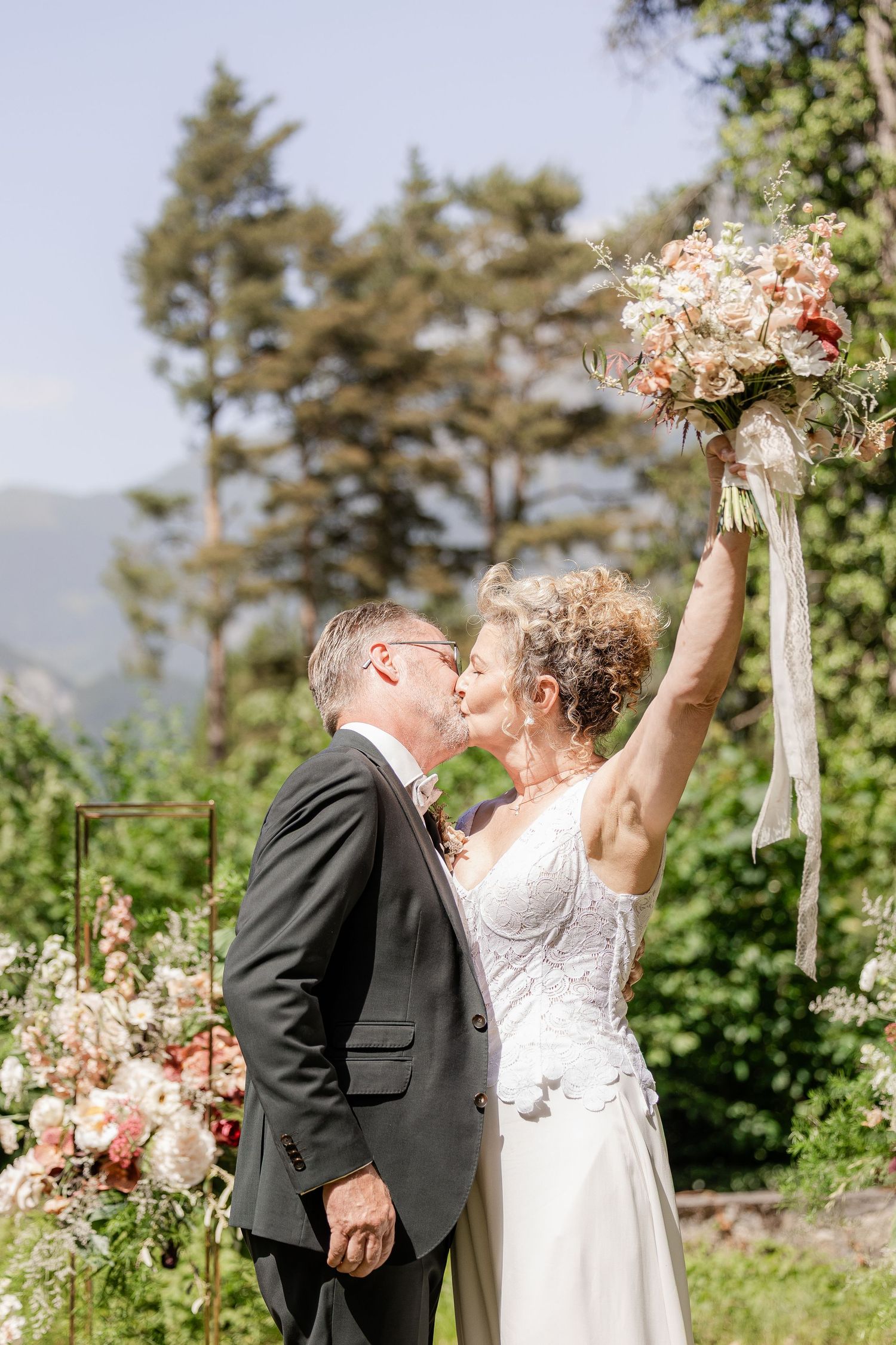 Mature bride raises bouquet while kissing groom outdoors at wedding ceremony with mountain backdrop.