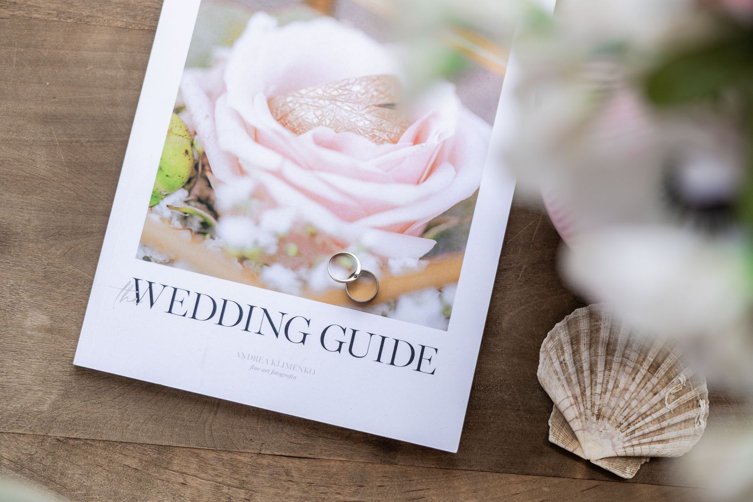 Wedding guide magazine with gold rings on pink rose cover, beside a seashell on rustic wood surface.