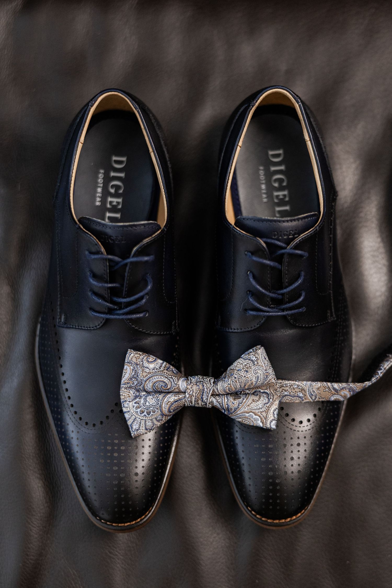 Black leather dress shoes with perforated details and a gray paisley bow tie placed on dark leather surface.