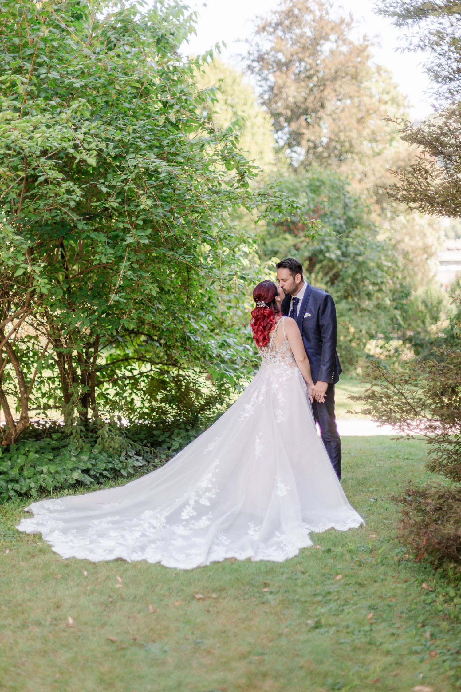 A couple in wedding attire pose together outdoors on a grassy lawn surrounded by lush green trees on their special day.