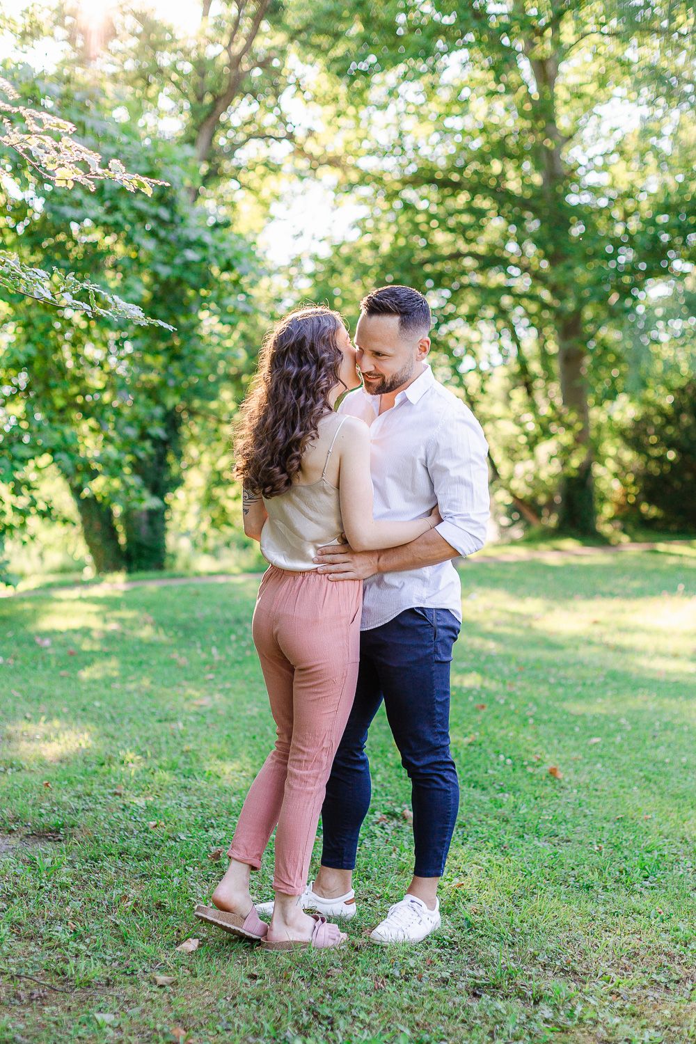 A couple shares a tender romantic moment together on a grassy hill surrounded by lush green trees in summer.