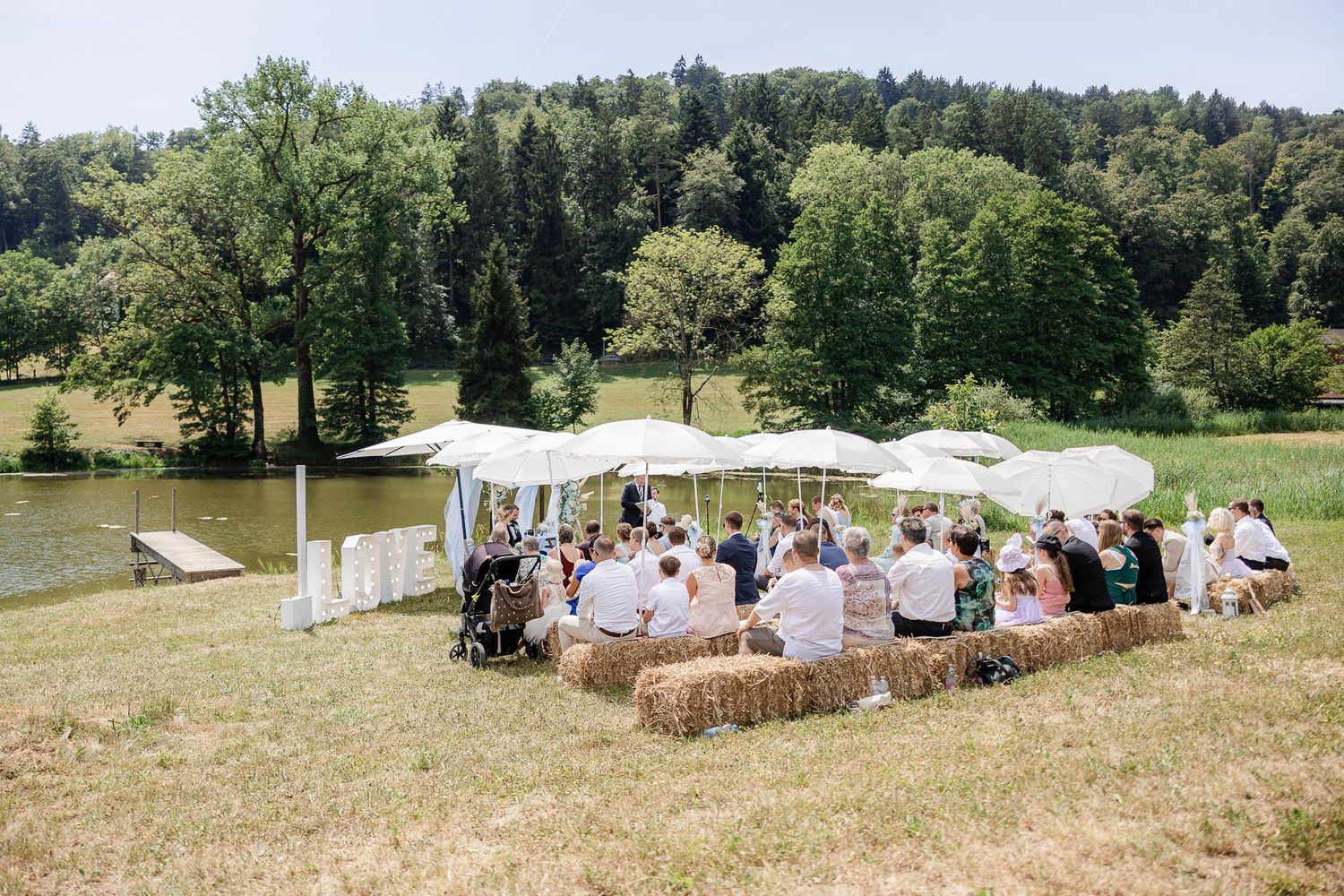 Outdoor wedding ceremony on a sunny day with guests seated on hay bales under a white canopy near a lake.