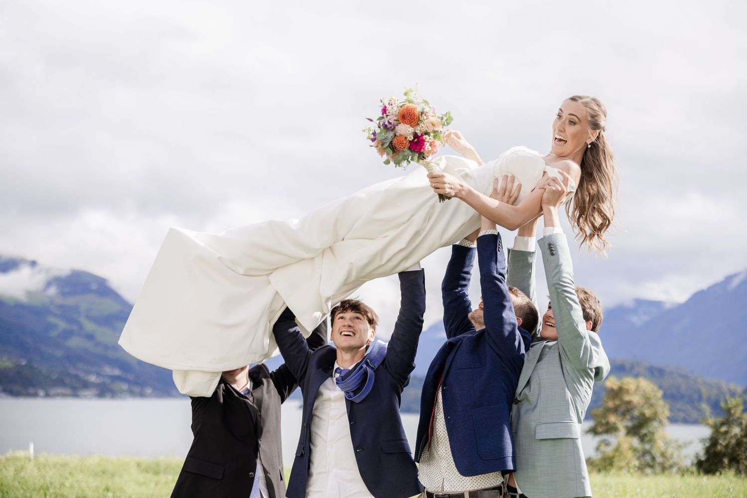 A group lifts a person in a flowing white dress against a mountain landscape backdrop.