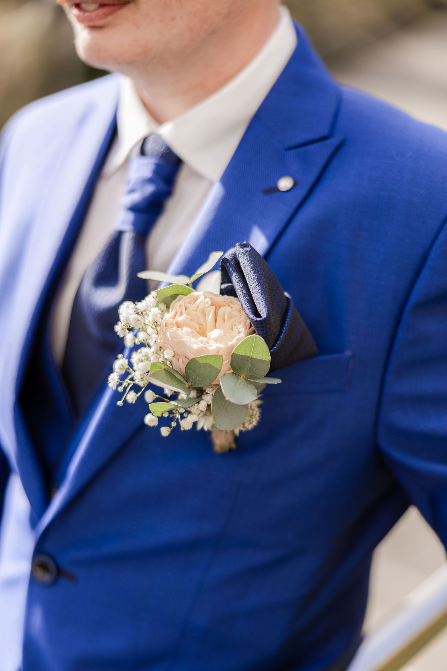 Groom in blue suit with boutonniere featuring peach rose, eucalyptus, and white flowers on lapel.
