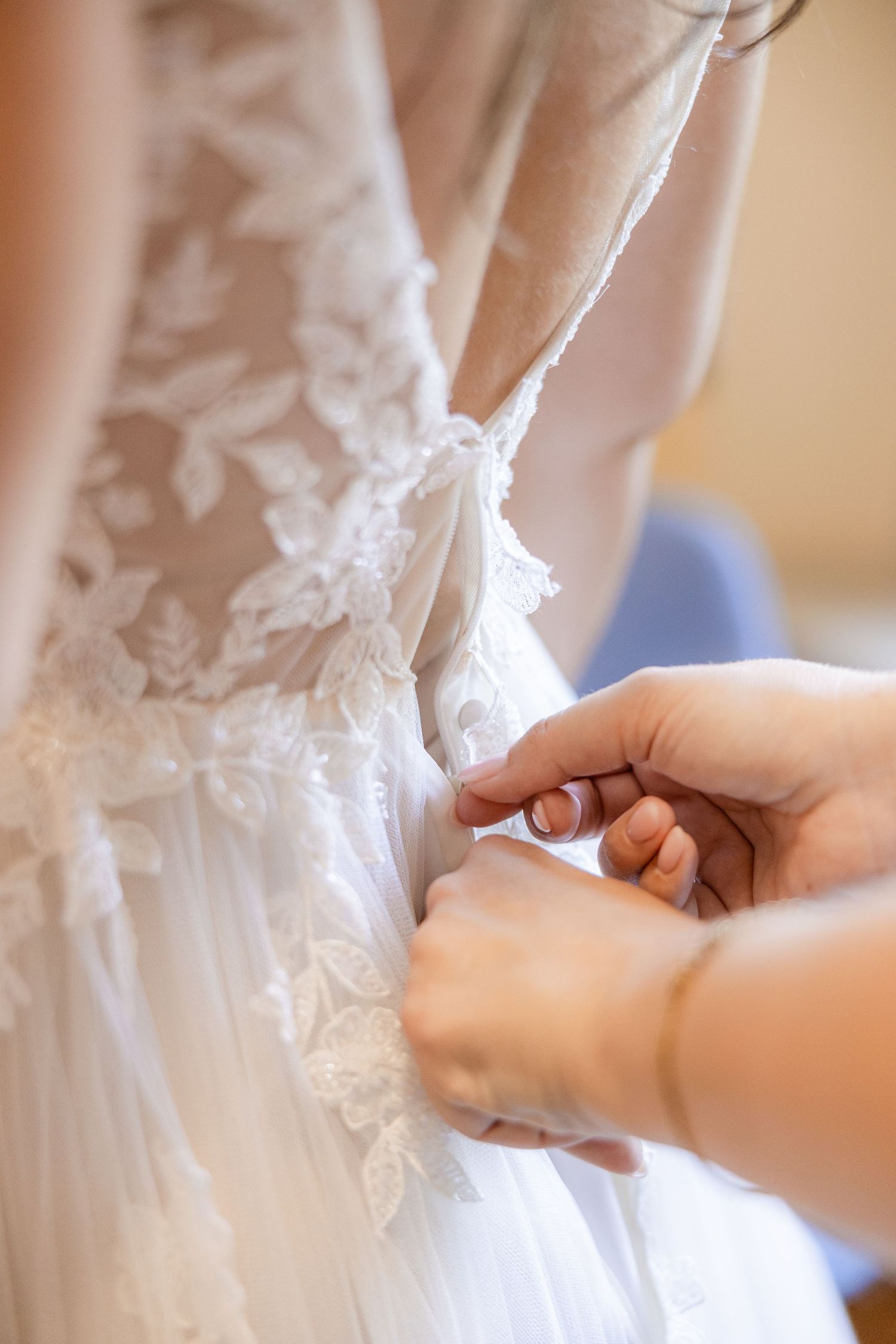 Hands fastening the back of a white lace wedding dress with floral applique details.