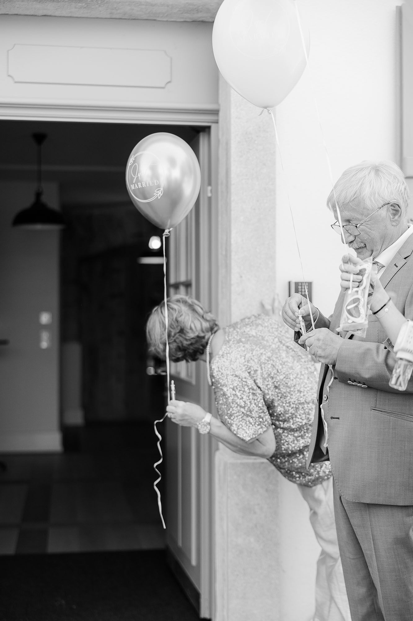 Elderly couple with balloons in black and white photo, man in suit assists woman at doorway during celebration.