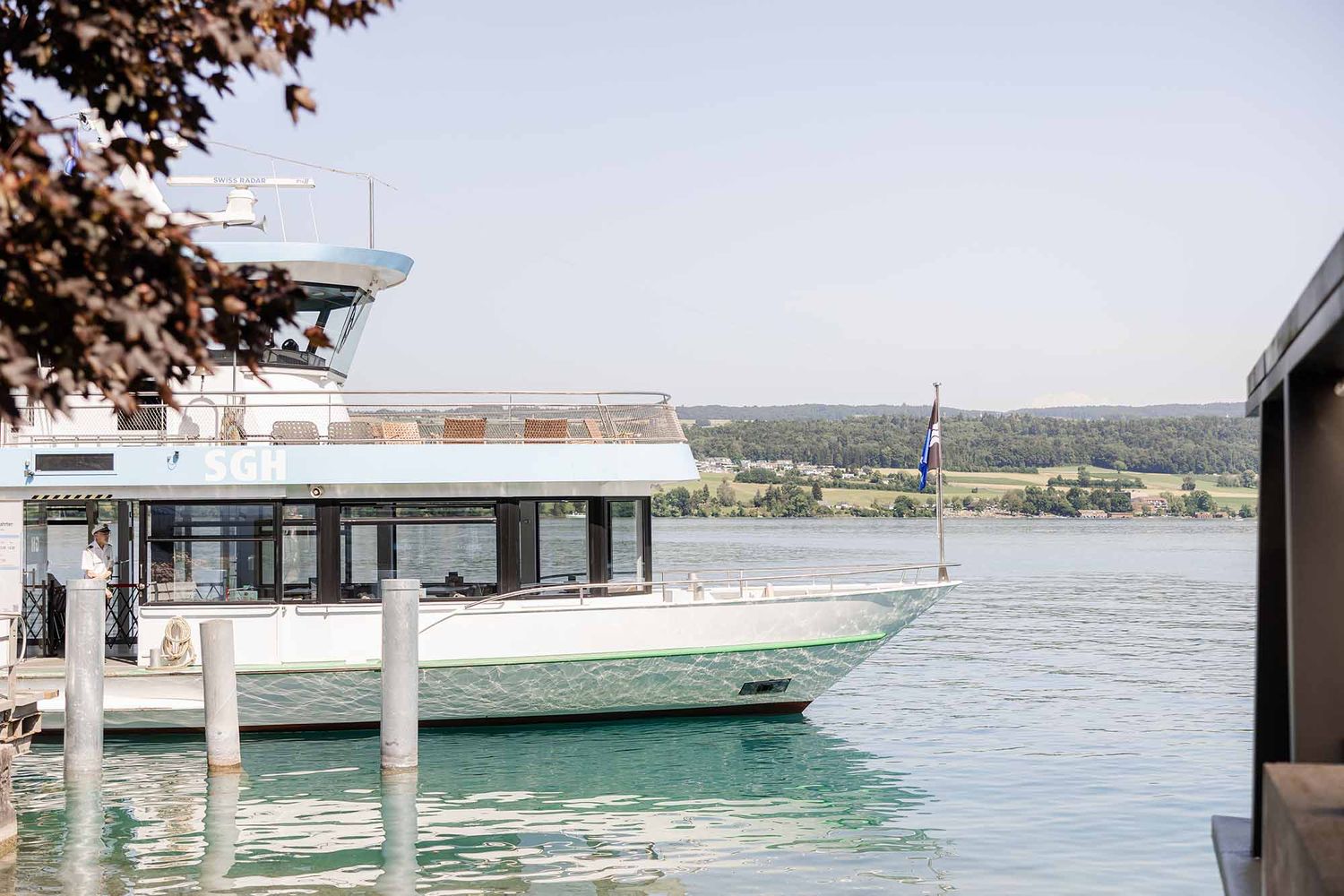 A luxury white motor yacht docked at a marina on calm blue waters with lush green trees in the background.