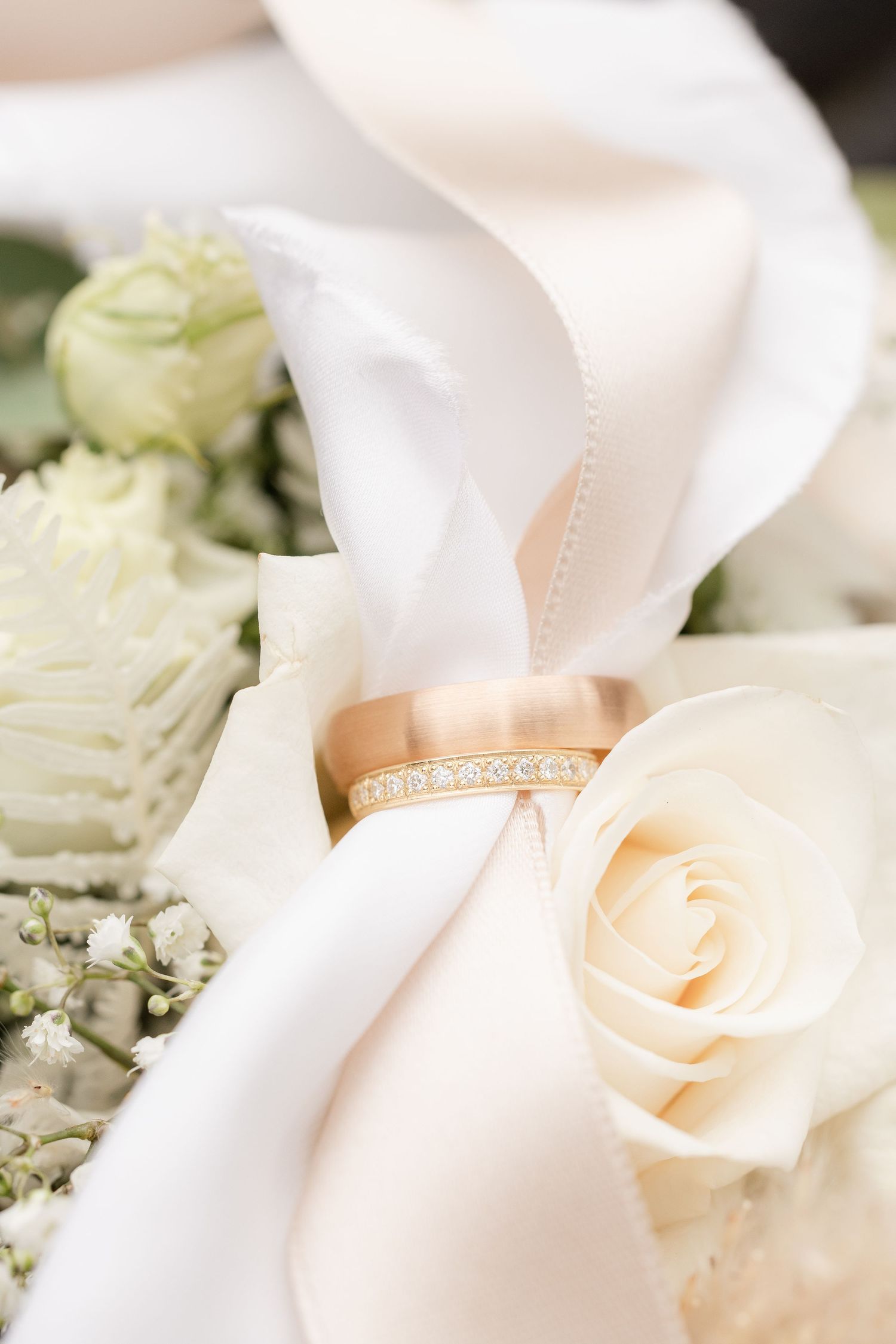 A diamond wedding band rests elegantly against white silk ribbon and cream roses in a romantic bridal detail shot. Greifensee, Zürich