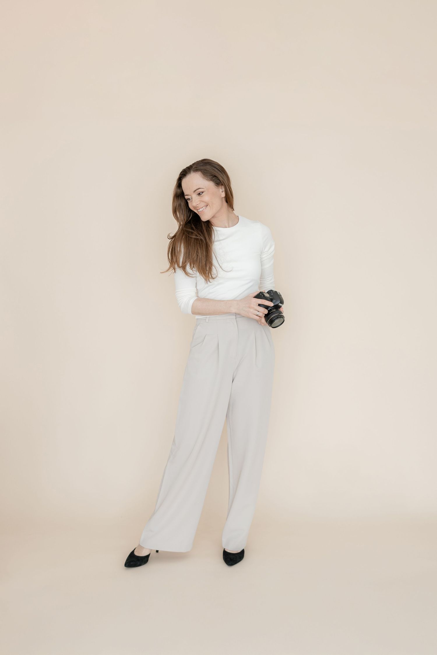 Person in casual business attire with white top and gray pants holds camera against neutral beige background.