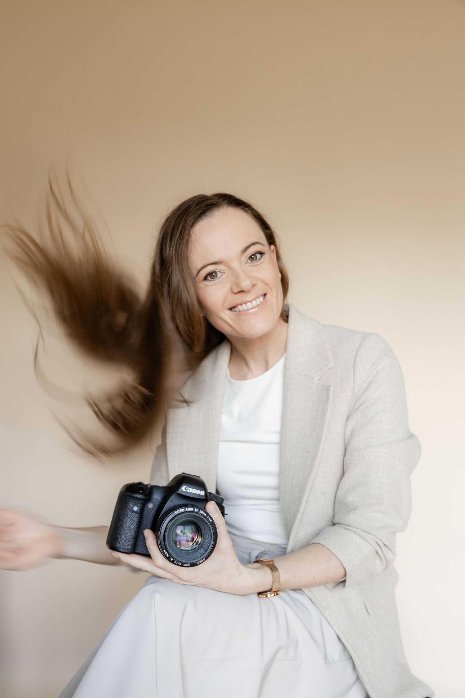 Professional photographer in light gray blazer and white dress holds DSLR camera while hair is caught in motion.