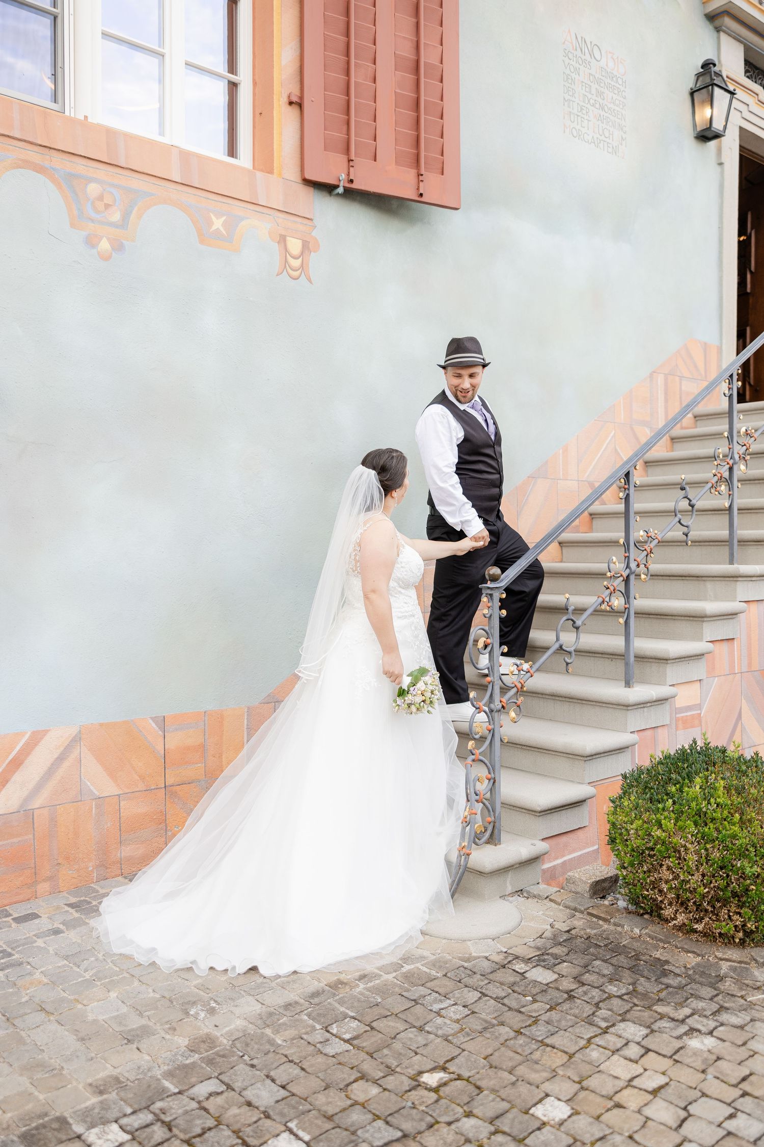 Couple in wedding attire pose on vintage stone steps by a painted wall with decorative architecture details.