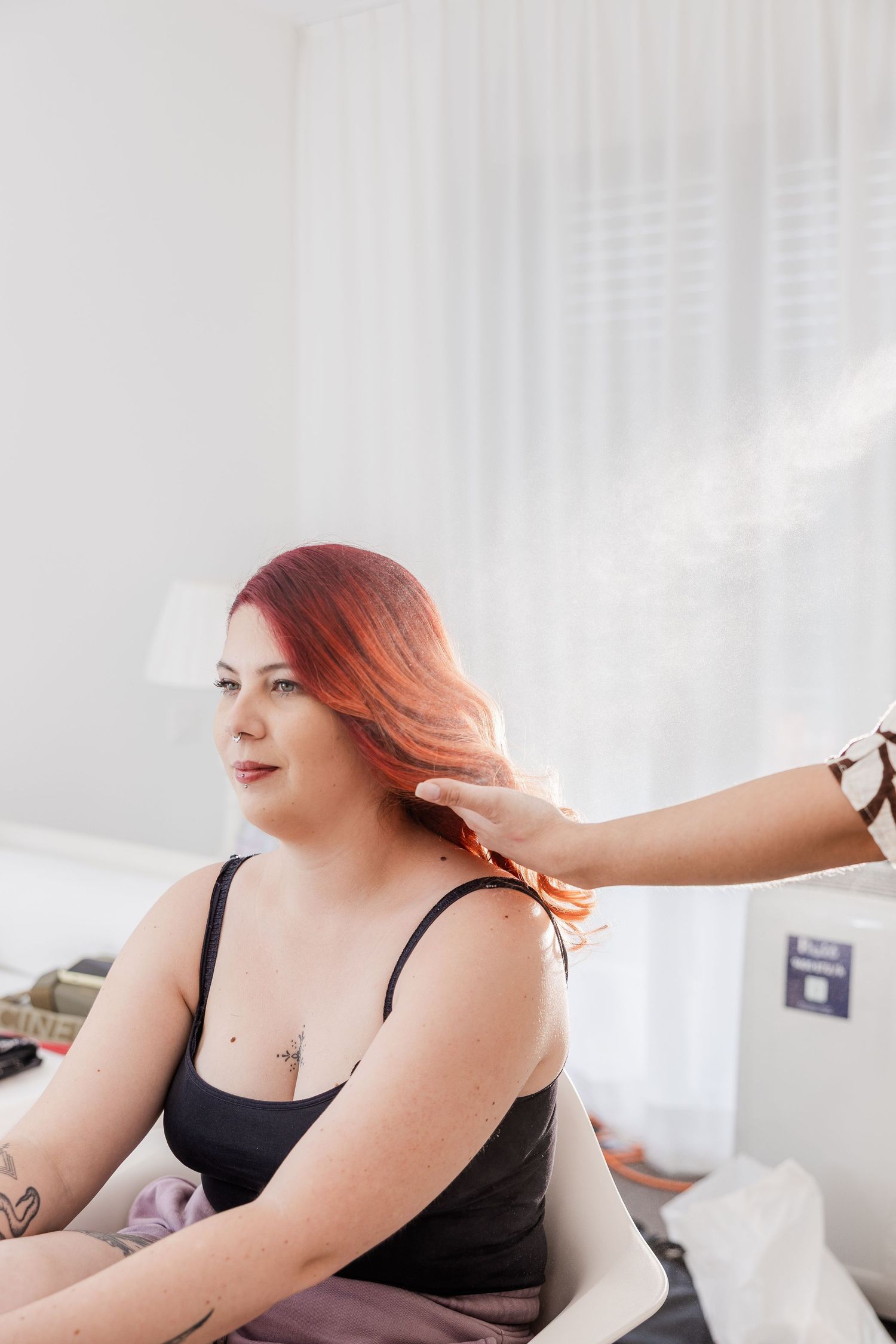 A smiling person with vibrant red hair wearing a black tank top sits casually in a bright, sunlit room.