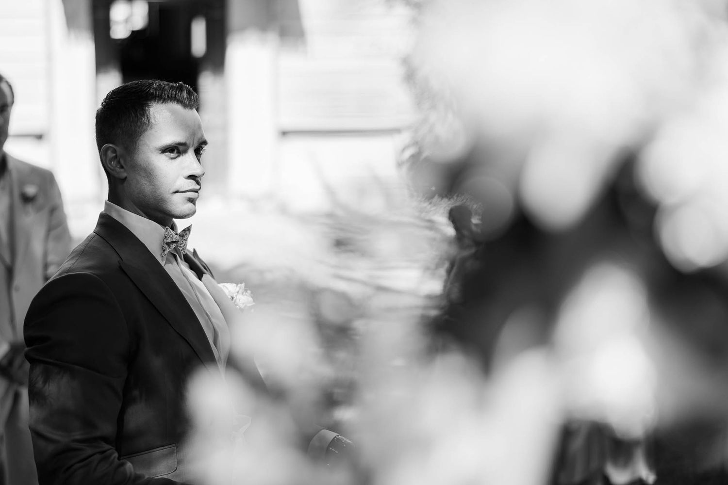 Black and white photo of a groom in a suit and bow tie waiting at the altar during a wedding ceremony.