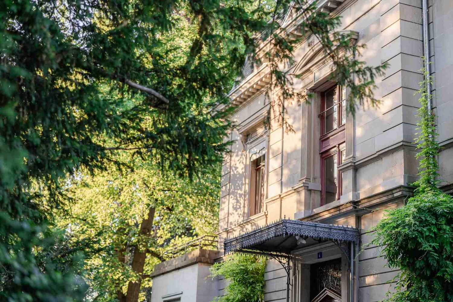Historic stone building facade surrounded by lush green trees with elegant entrance canopy on sunny day.