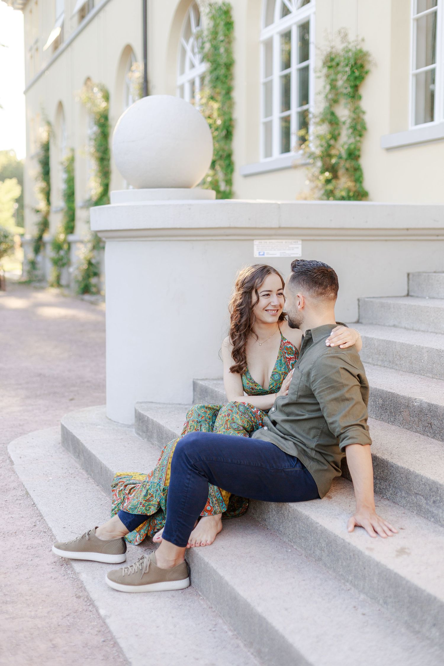A couple sits close together on elegant stone steps outside a building with white arched windows and climbing vines.