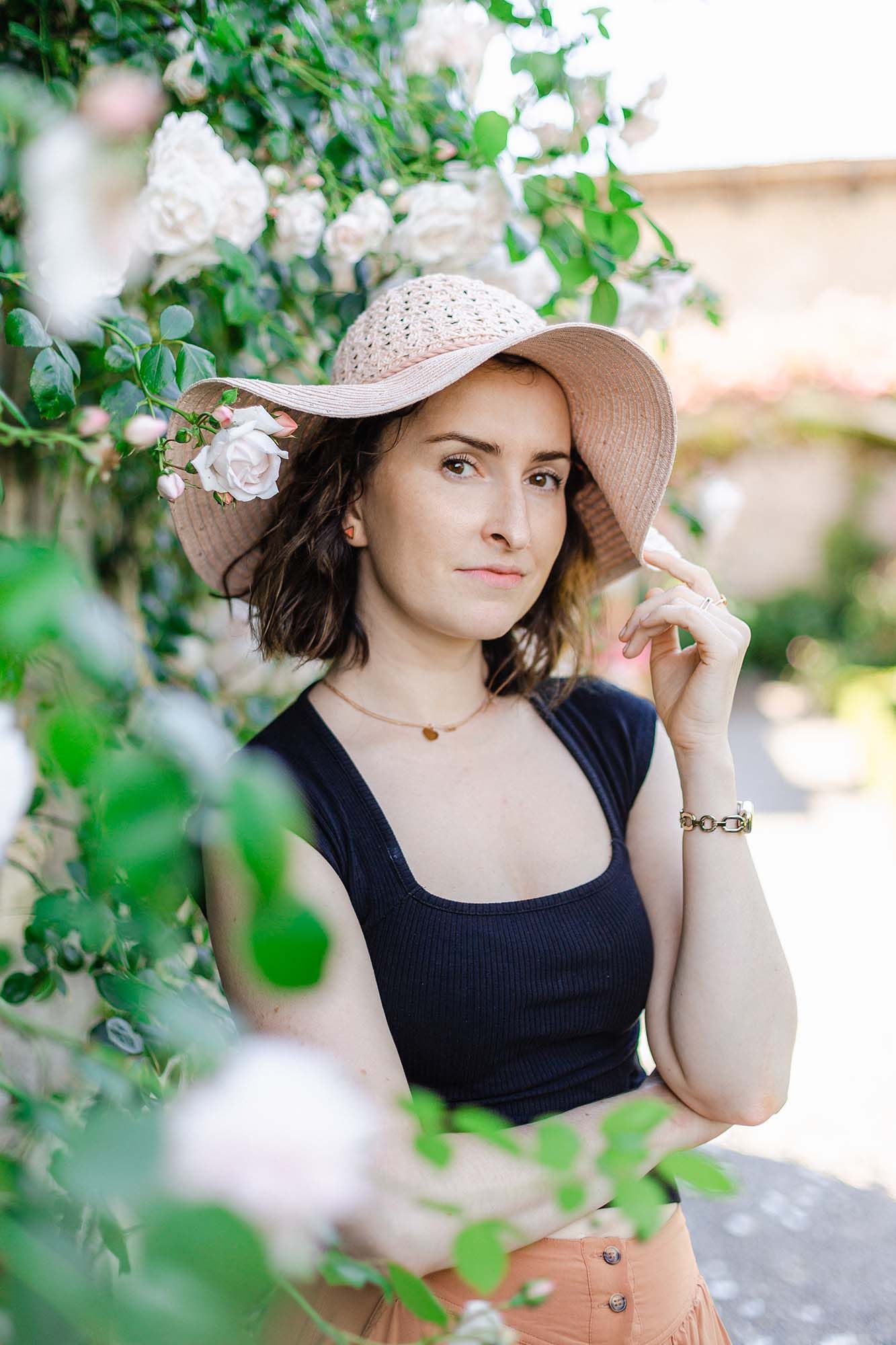 Woman in pink sun hat and black tank top poses among white roses in a lush garden setting.