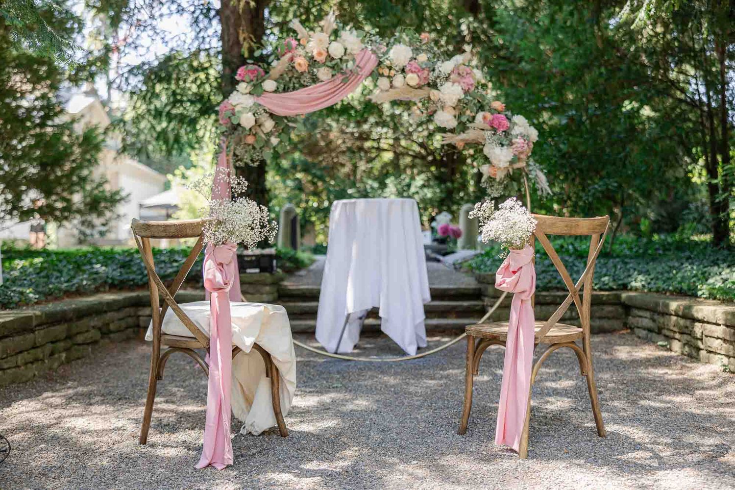Romantic outdoor wedding ceremony setup with floral arch, pink ribbon chairs, and white draped table amid lush greenery.