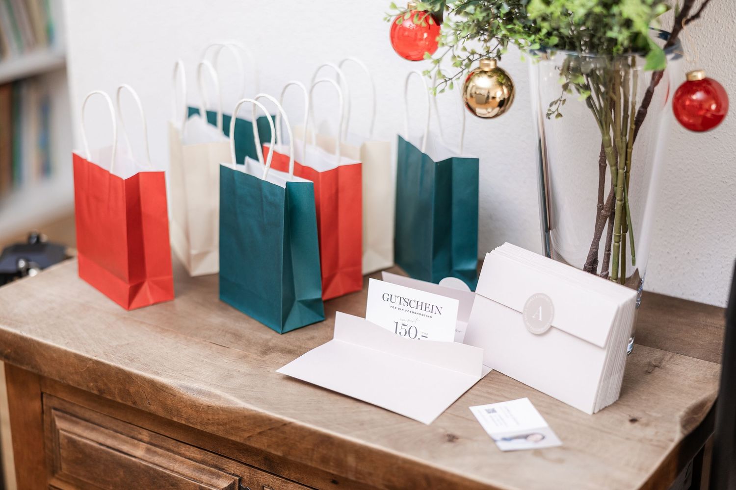 Holiday gift bags in red and green sit on a wooden table with greeting cards and festive ornaments hanging above.