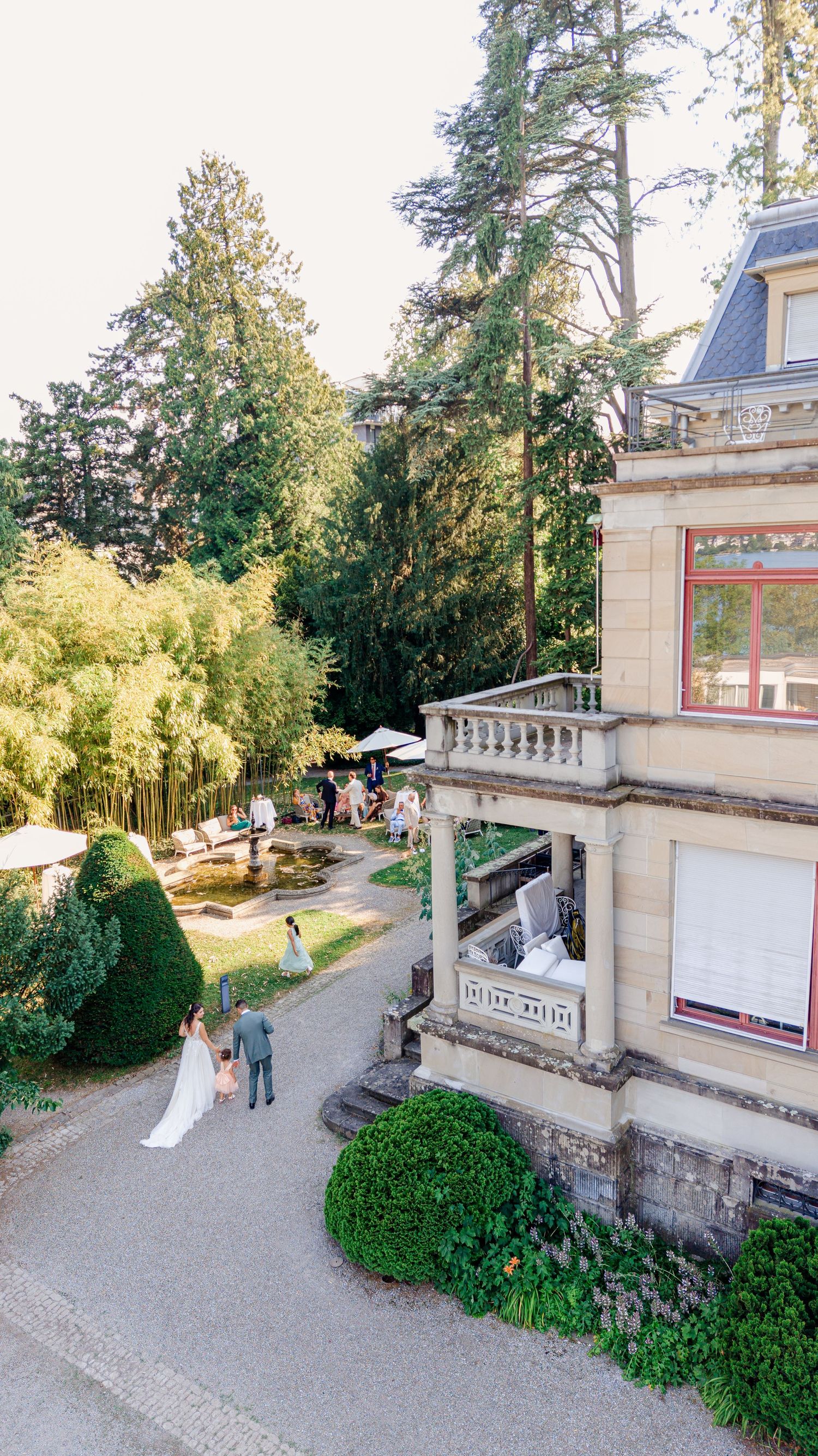 Aerial view of a bride and groom walking beside an elegant stone mansion with lush garden, fountain, and tall trees.
