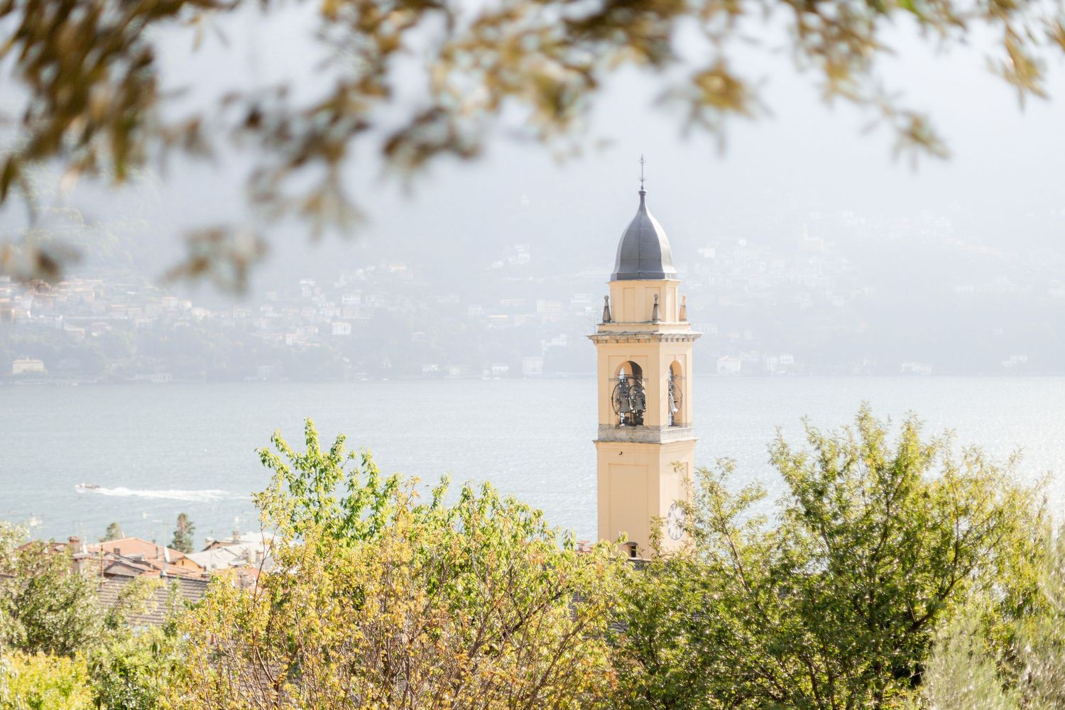 Church bell tower rises above green foliage with a misty Mediterranean landscape in the background.