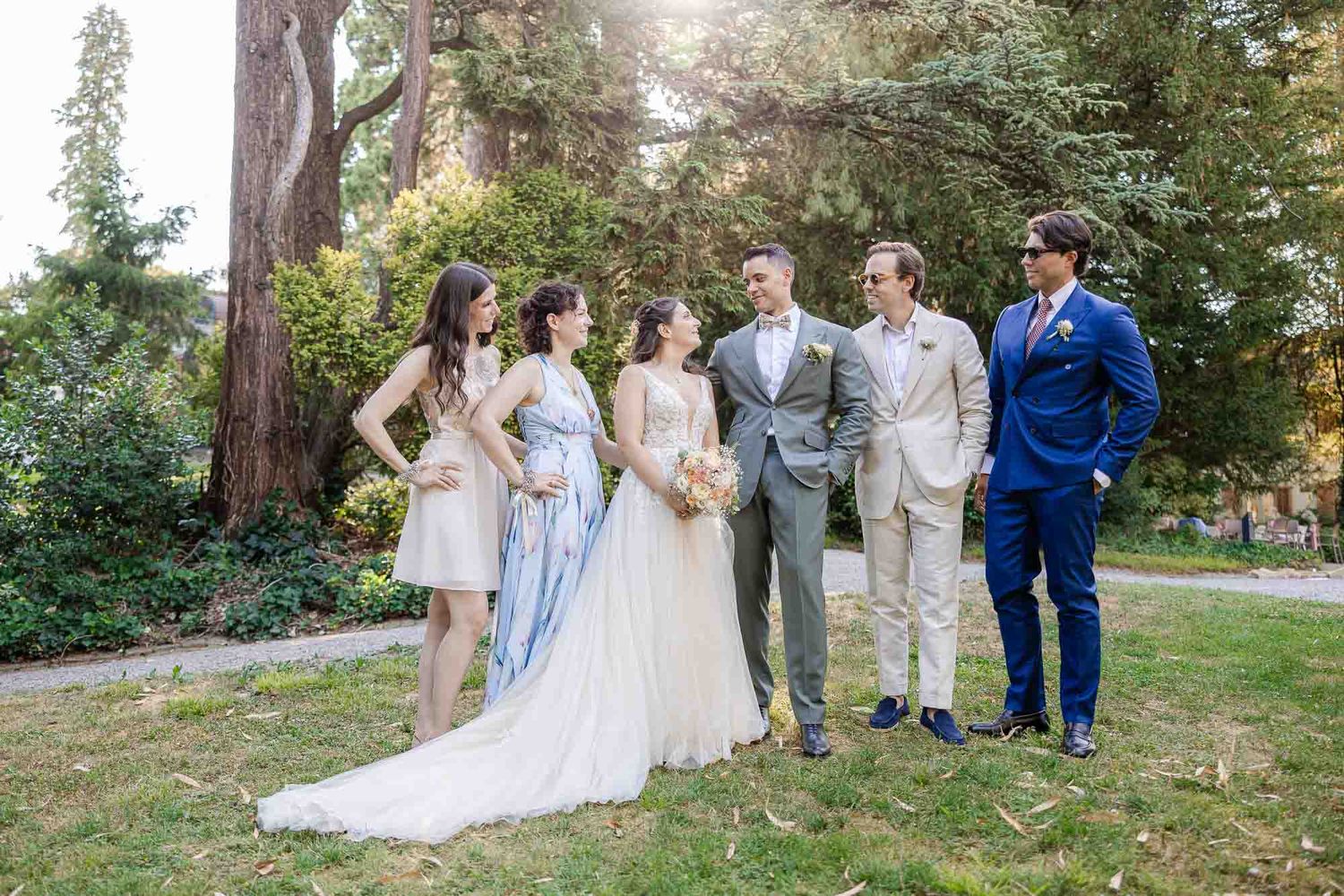 Wedding party posing outdoors in elegant attire, bride in white gown with bridesmaids and groomsmen among lush green trees.