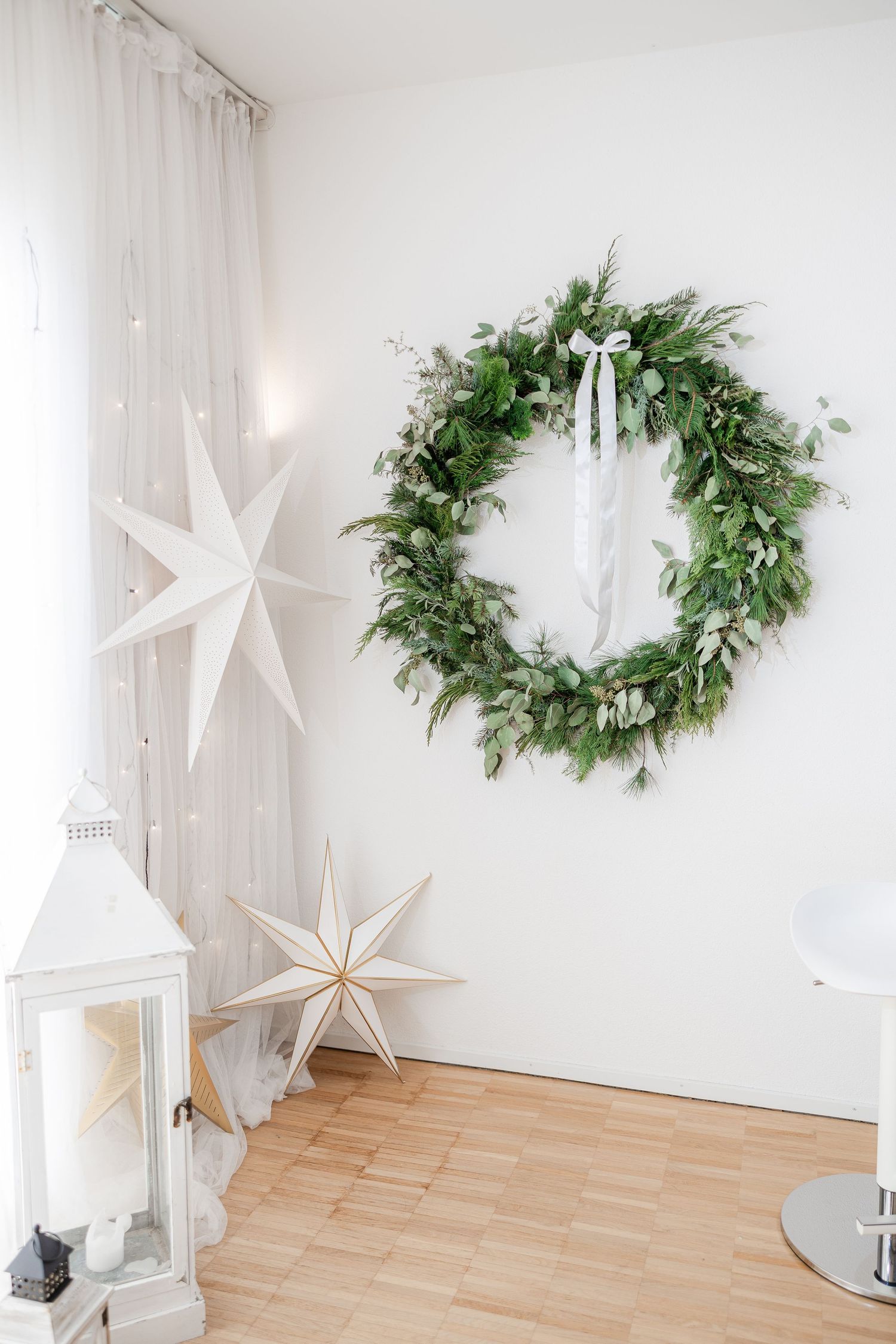 Minimalist Christmas decor featuring a fresh evergreen wreath, white paper stars and lantern against white walls.