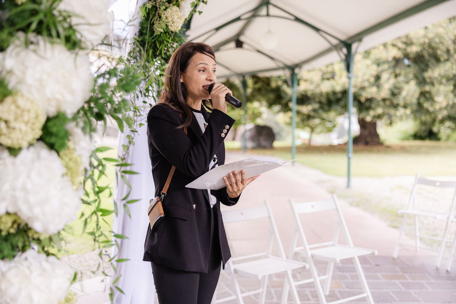 A wedding officiant in black attire speaks at an outdoor ceremony under a white tent with floral decorations.