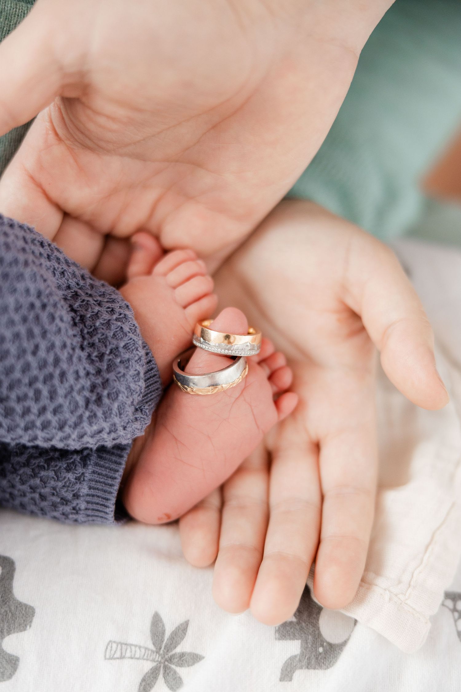 Two adult hands cradling a newborn baby's tiny feet with gold and silver wedding rings placed on the infant's toes.