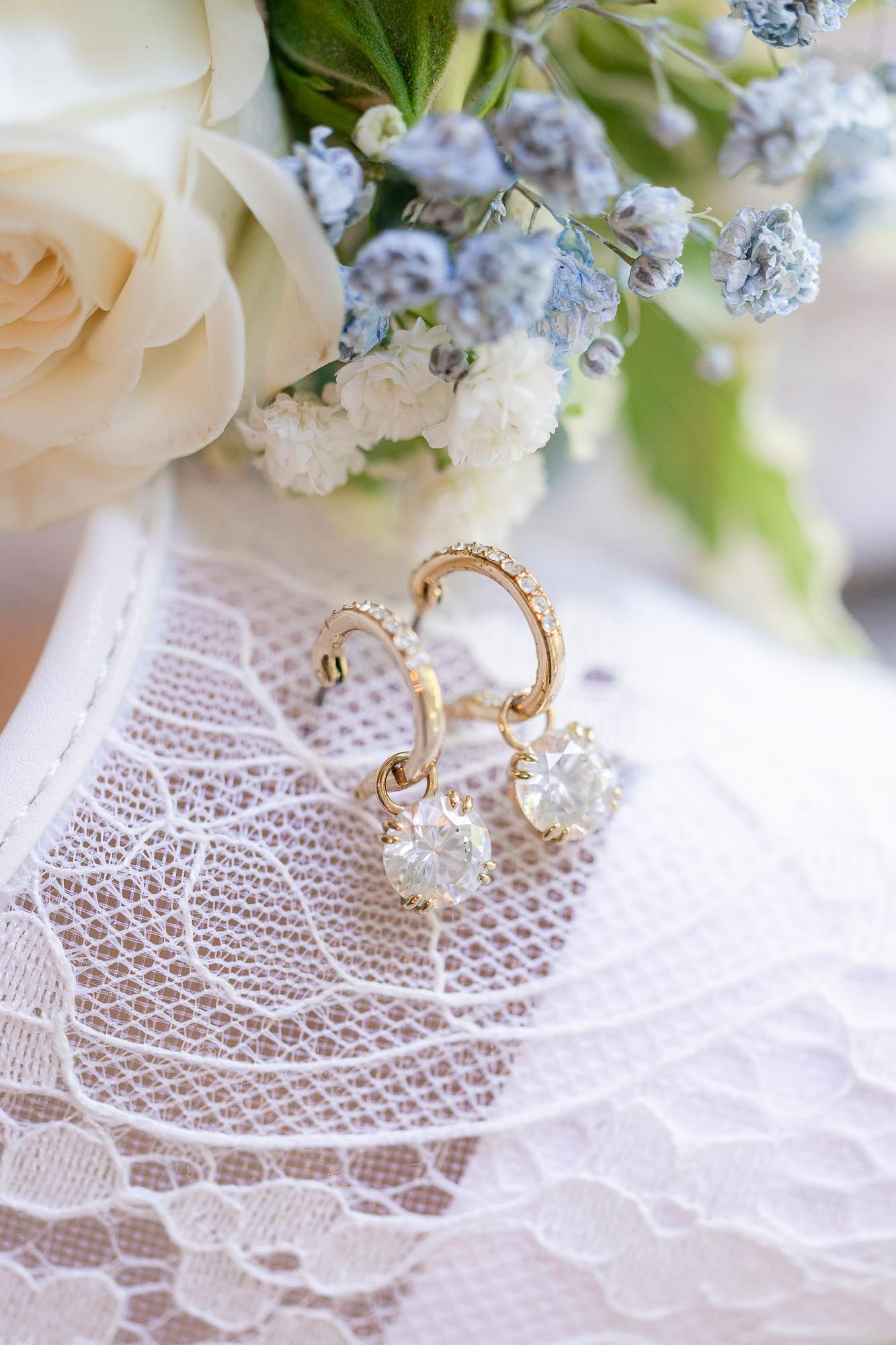 Gold hoop earrings with diamond drops on white lace fabric near roses and blue flowers.