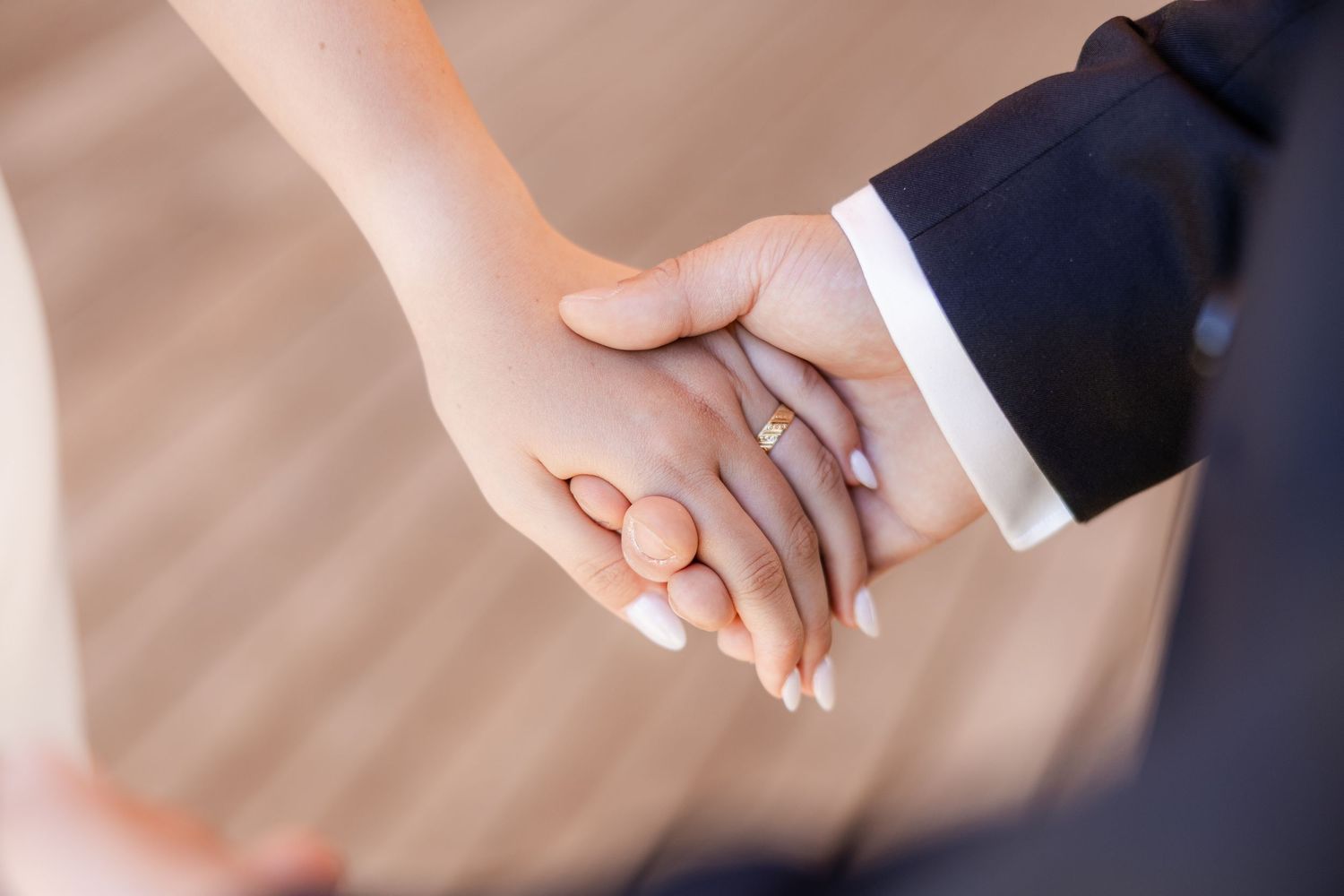 Close-up of a couple holding hands, bride wearing an engagement ring, groom in a dark suit at a wedding ceremony.