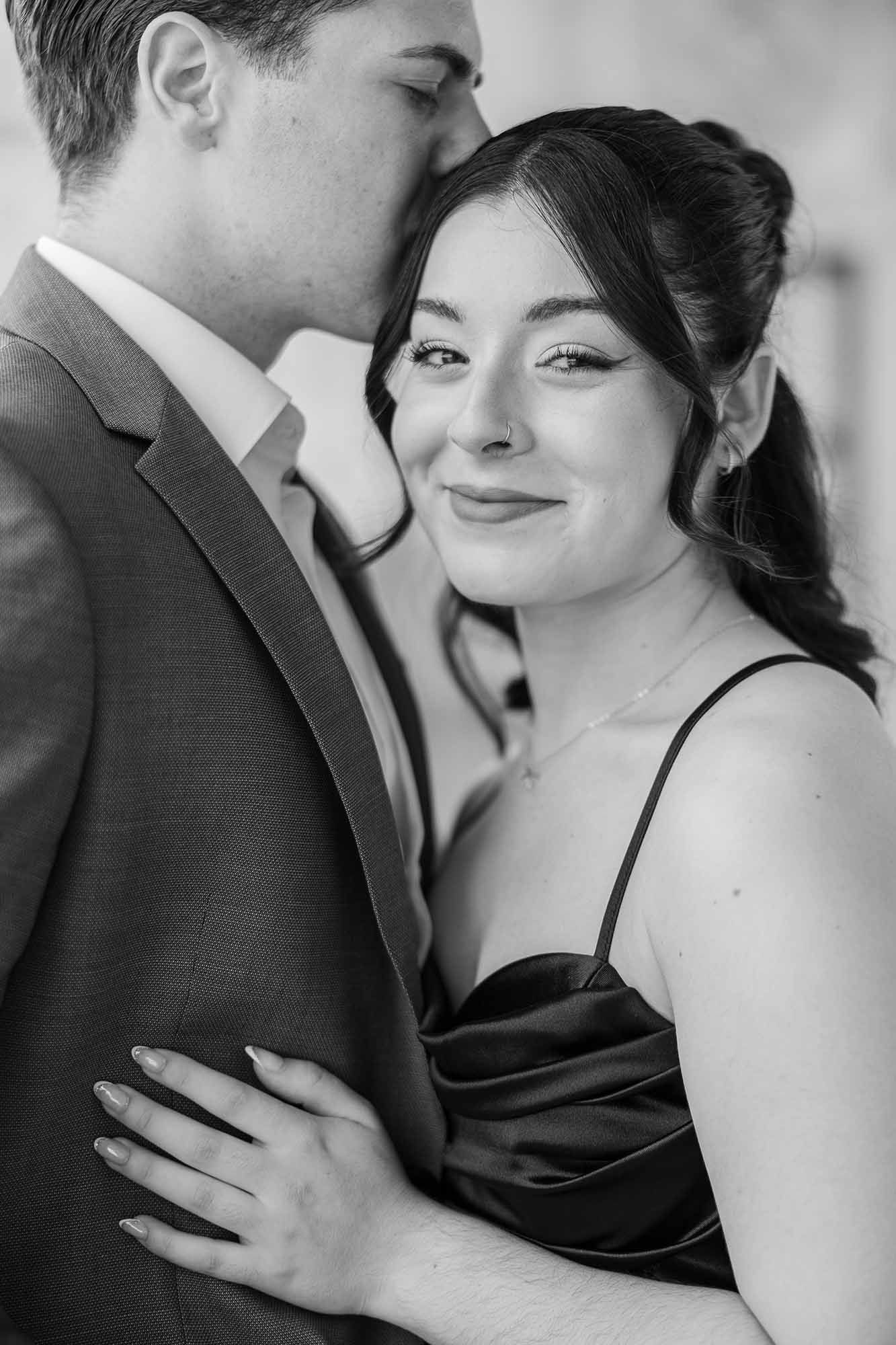 Black and white photo of a couple embracing, woman smiling at camera wearing satin dress, man in suit kissing her cheek.