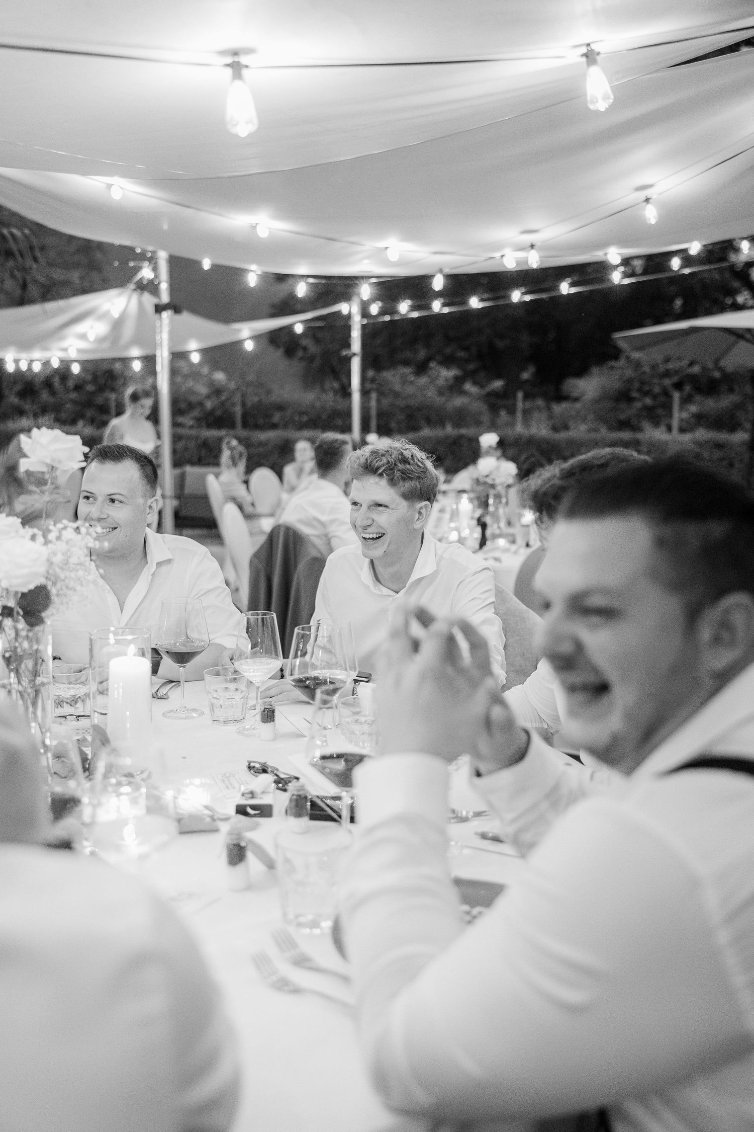 Wedding guests laugh and celebrate together at reception tables under string lights during an evening outdoor celebration.