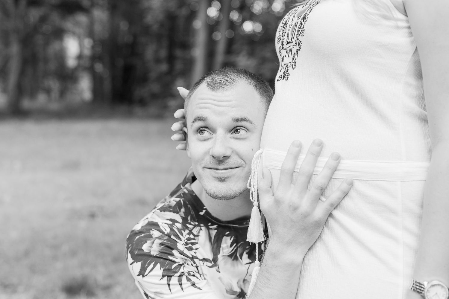Black and white maternity photo showing an excited expression while listening to pregnant belly outdoors - Schwangerschaftsfoto Aargau