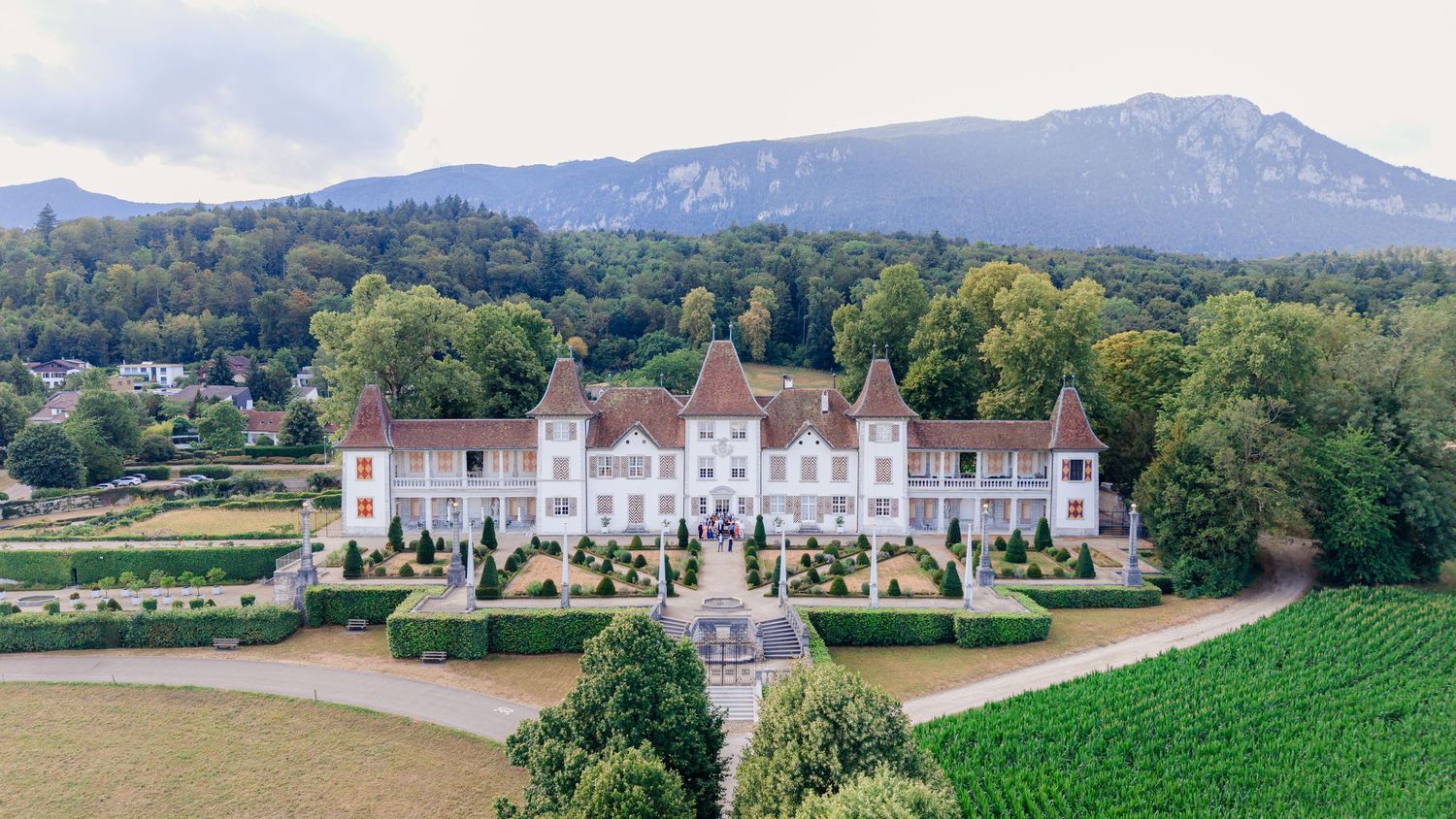 Aerial view of a grand French château with manicured gardens, vineyards, and mountain backdrop.