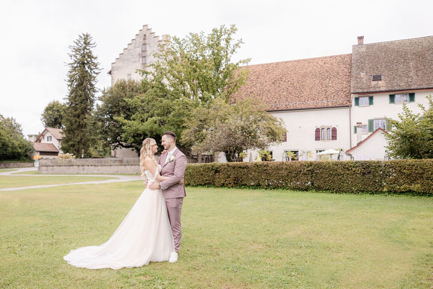 Two people share a romantic embrace on a lush green lawn with rustic buildings and trees in the background.